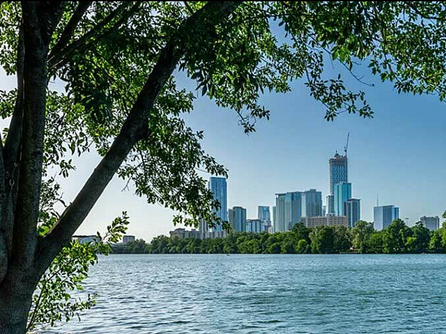 Scenic view of a serene lake lined with lush trees and a bustling city skyline in the background, offering a blend of natural tranquility and urban sophistication.