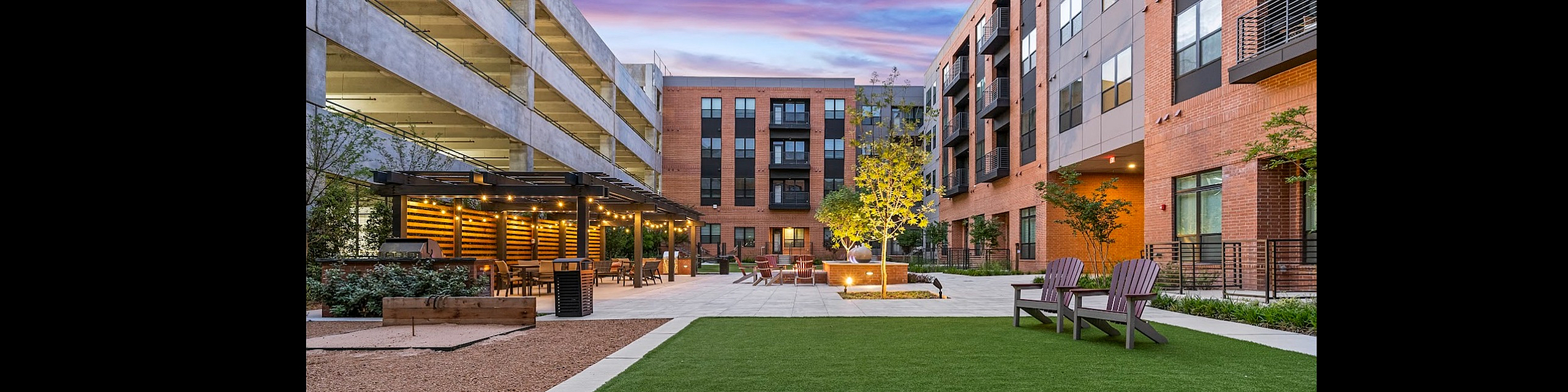 Charming courtyard with cozy seating, pergola, and vibrant landscaping at a modern apartment complex, under a stunning sunset sky.