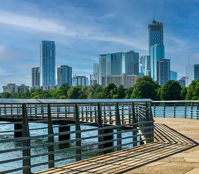 Scenic waterfront view with a modern skyline, featuring a boardwalk overlooking a serene river and lush greenery.