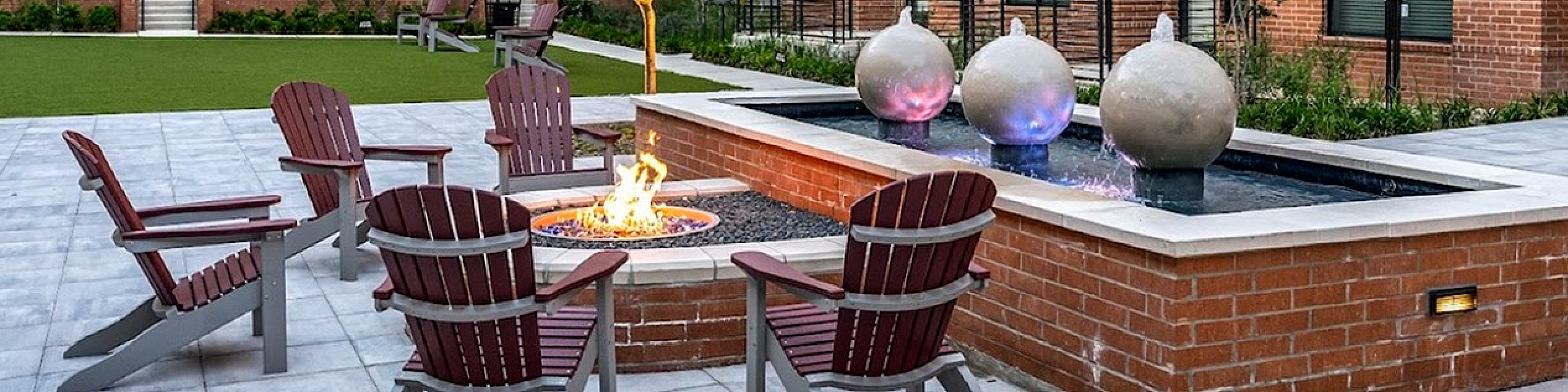 Inviting courtyard with Adirondack chairs around a cozy fire pit, modern fountain, lush greenery, and stylish brick apartment backdrop.