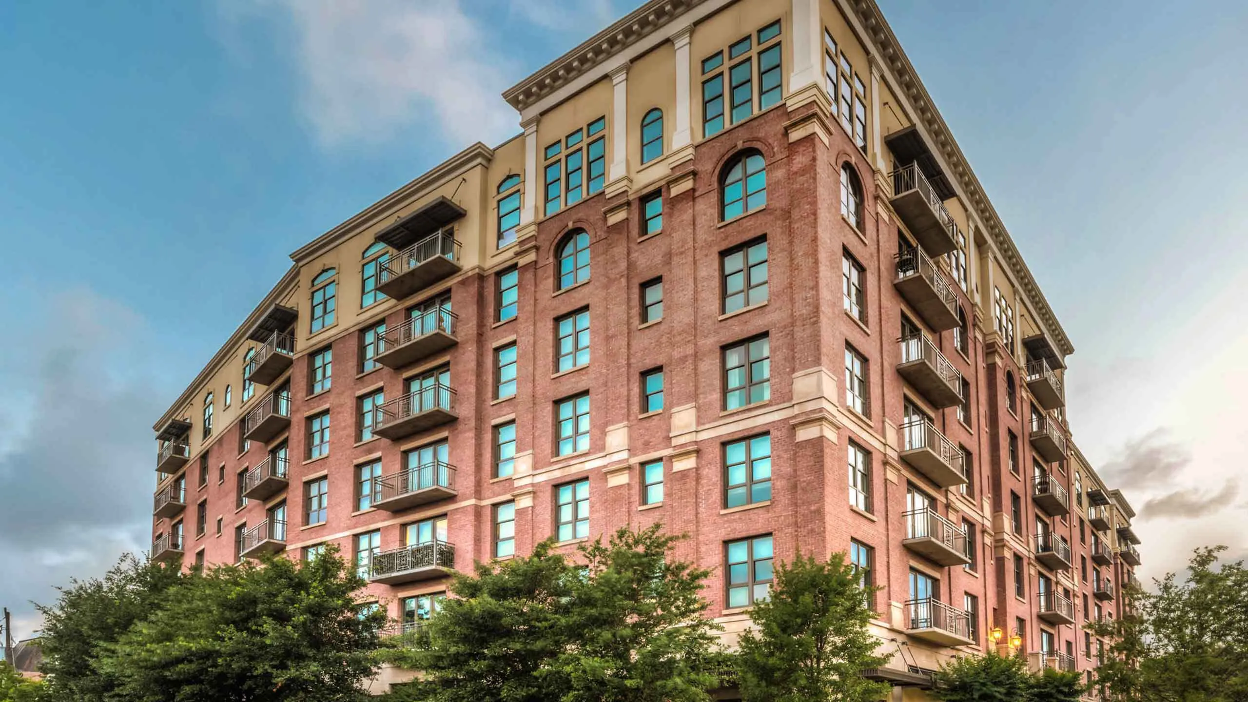 Elegant brick apartment building with modern balconies and large windows, surrounded by lush greenery under a clear blue sky.