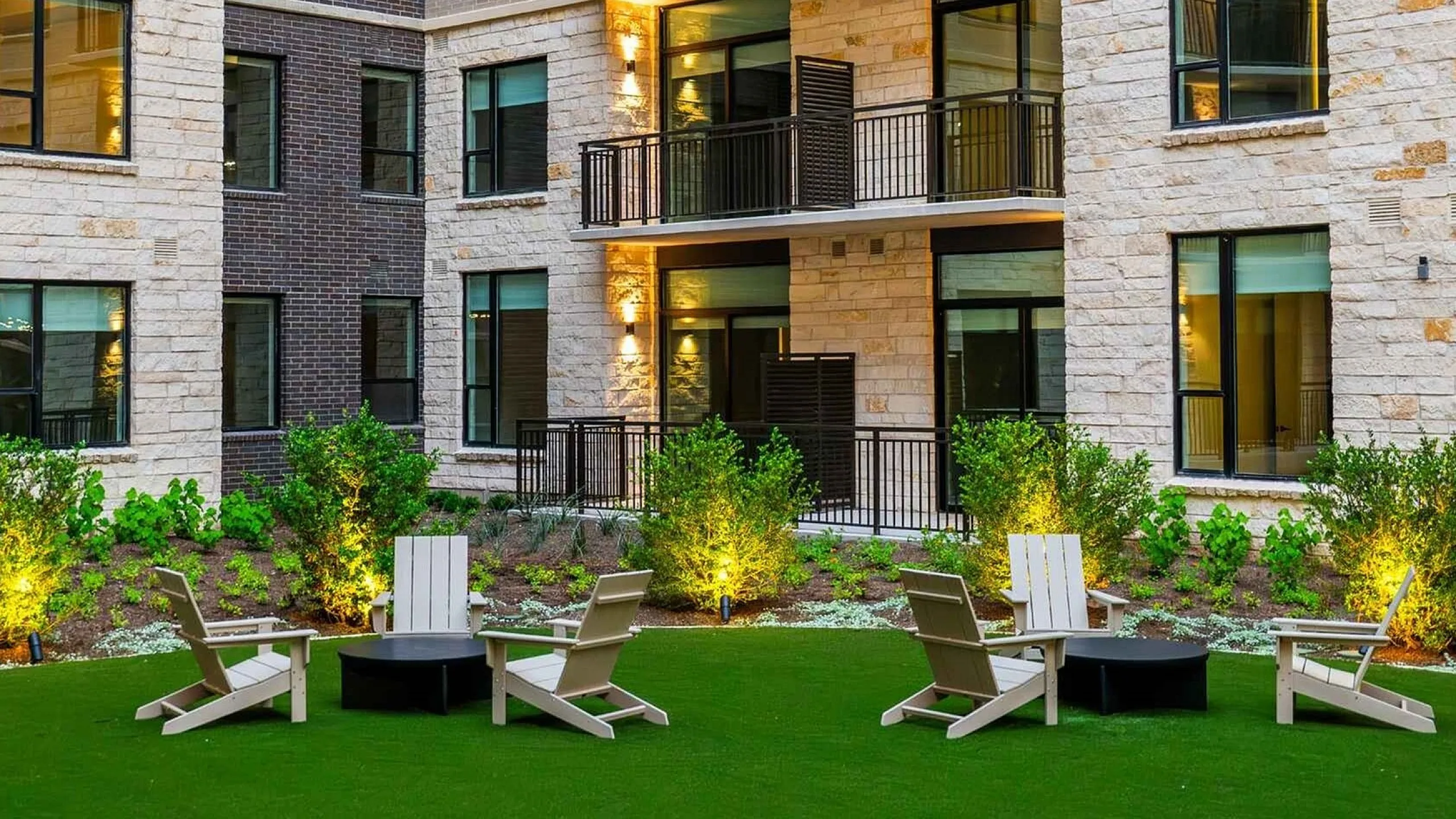 Modern apartment complex with illuminated balconies, lush green courtyard, and inviting seating area for relaxation.