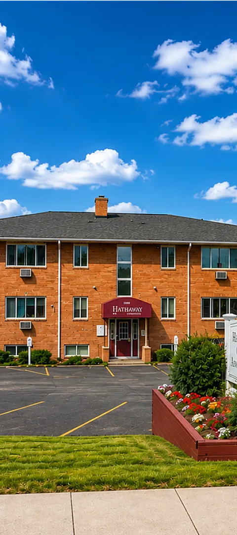Charming brick apartment building at Hathaway Court with lush landscaping, ample parking, and picturesque blue skies.
