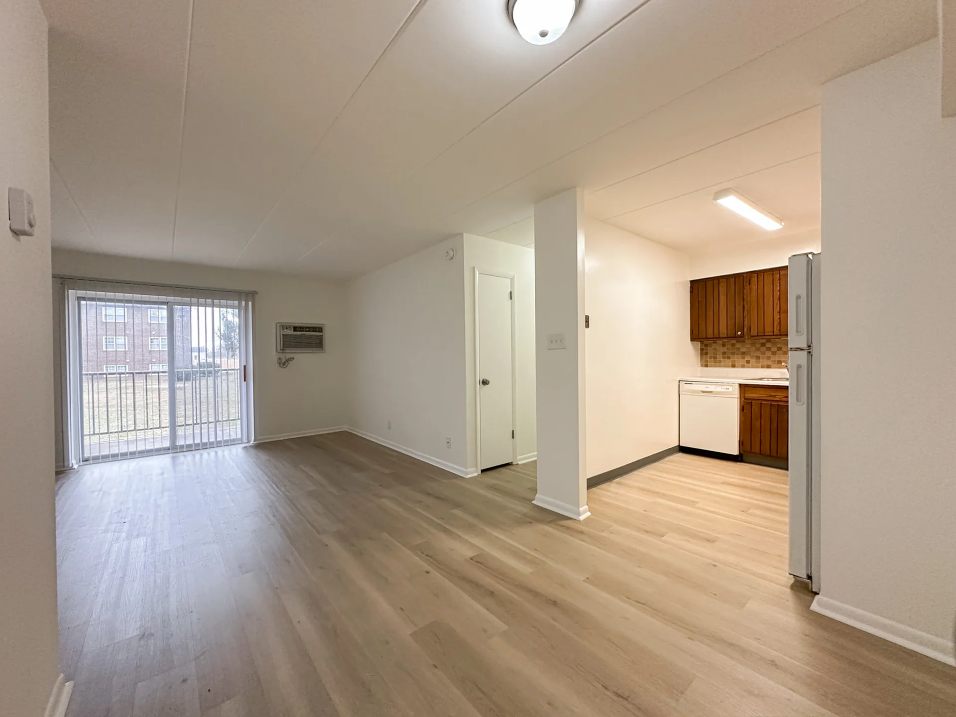 Bright and airy apartment interior featuring an open-concept living area with modern laminate flooring, sliding glass doors to a balcony, and an adjacent cozy kitchen with wooden cabinets.