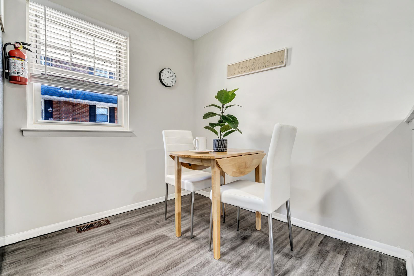 Cozy dining nook with a modern wooden table, two white chairs, and a potted plant, complemented by a bright window and sleek decor.