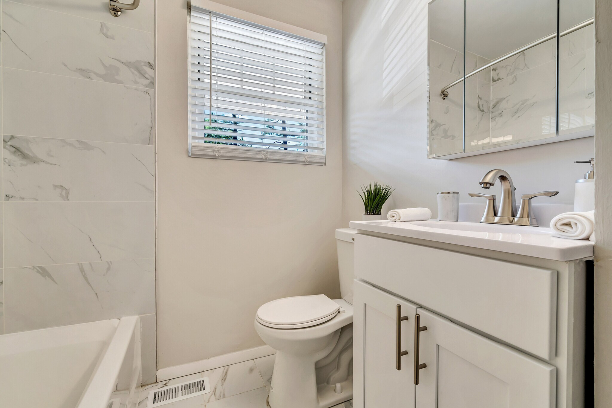 Modern bathroom adorned with sleek marble tiles, featuring a stylish vanity, polished chrome fixtures, and a sunlit window with blinds.