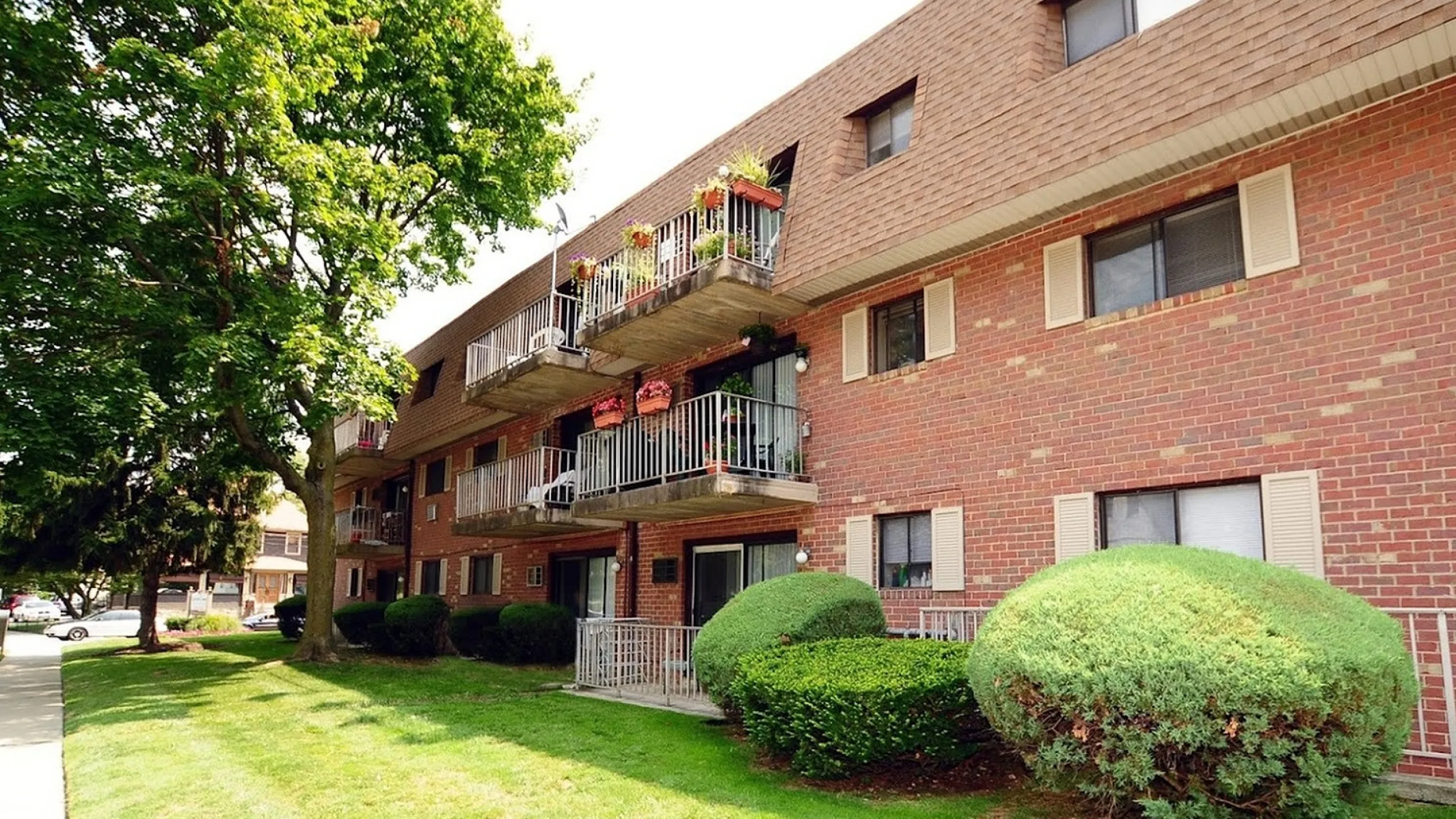 Beautiful brick apartment building with balconies adorned with flowers, surrounded by lush greenery.