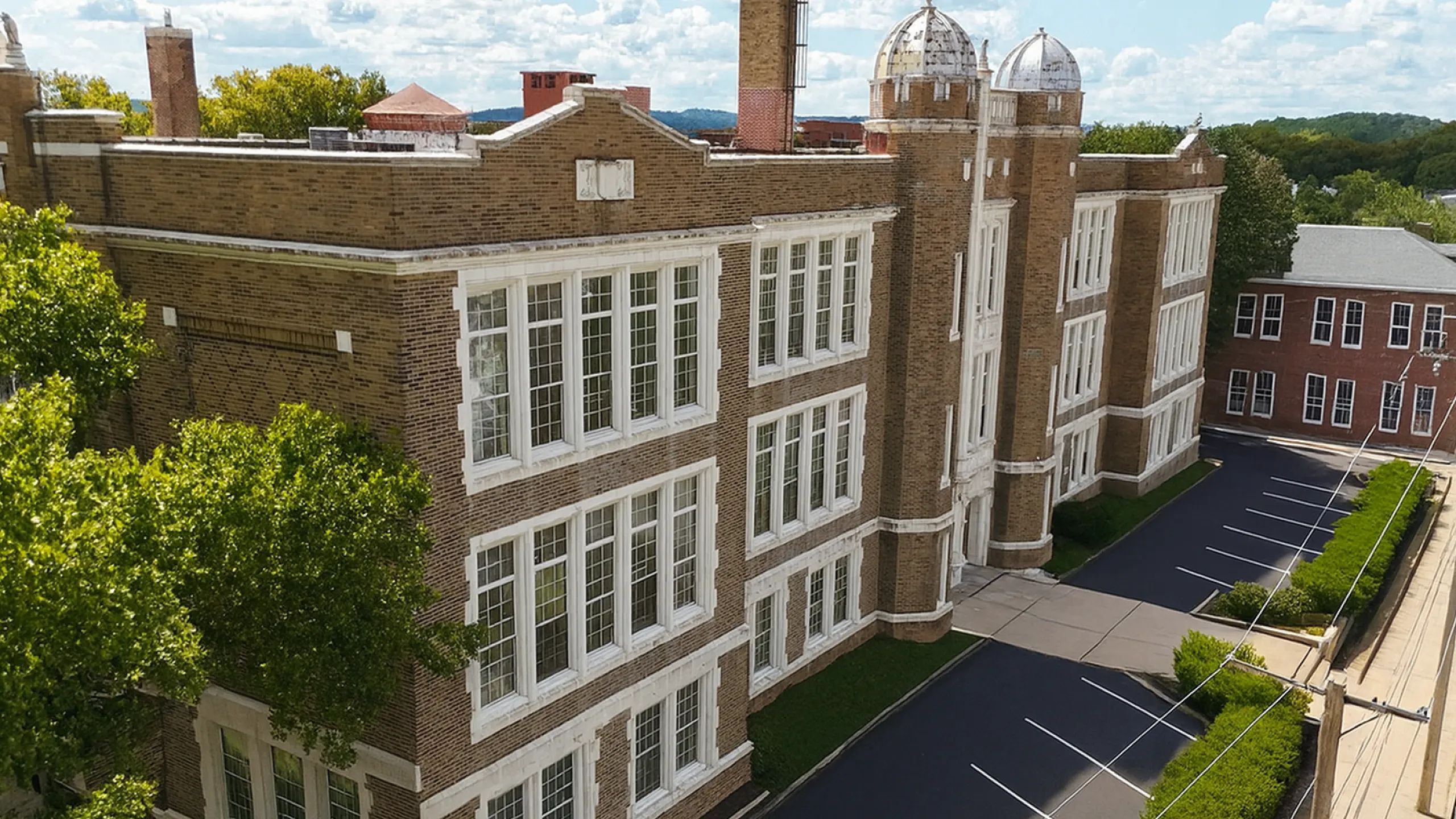 Charming historic building with classic architecture, large windows, and spacious parking, surrounded by lush greenery on a bright sunny day.