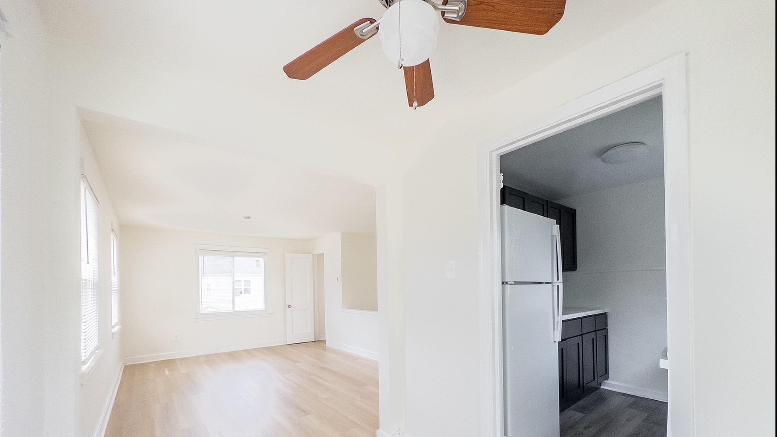 Bright and airy living room with light wood flooring and a stylish ceiling fan, leading into a sleek, modern kitchen with dark cabinetry.