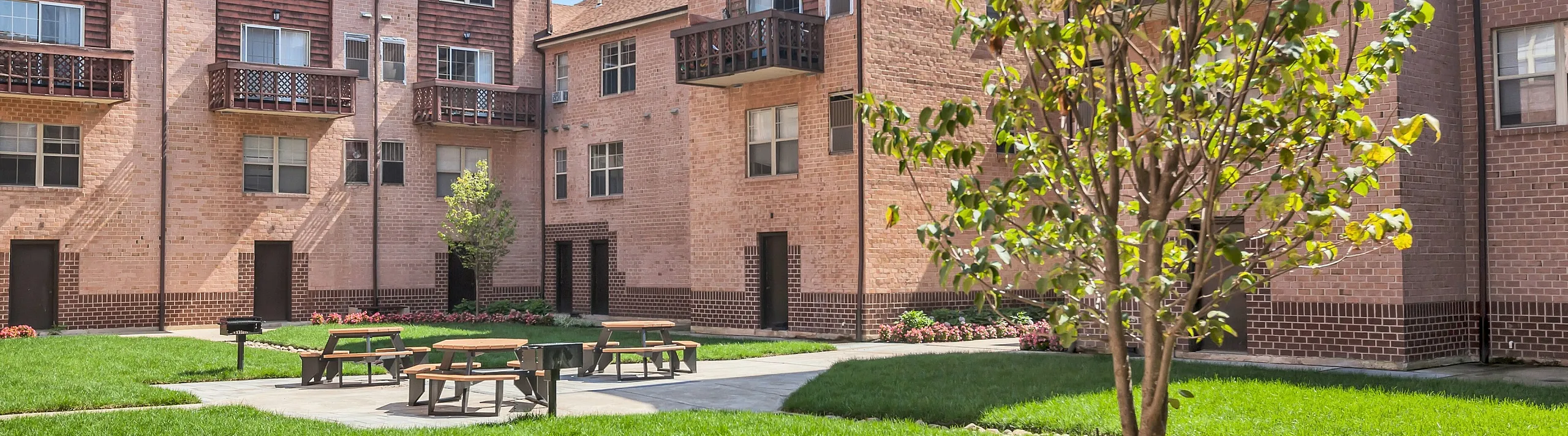 Beautiful brick apartment complex with lush green courtyard, picnic tables, and vibrant flower beds, offering a serene living environment.