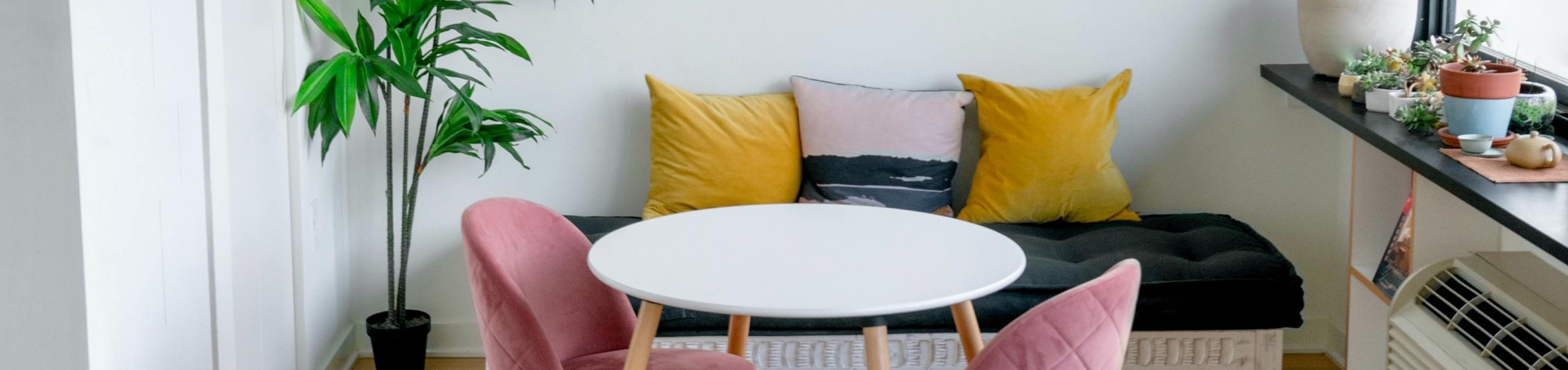 Charming apartment nook with cozy bench, vibrant cushions, sleek round table, and modern pink chairs, perfect for relaxation and dining.