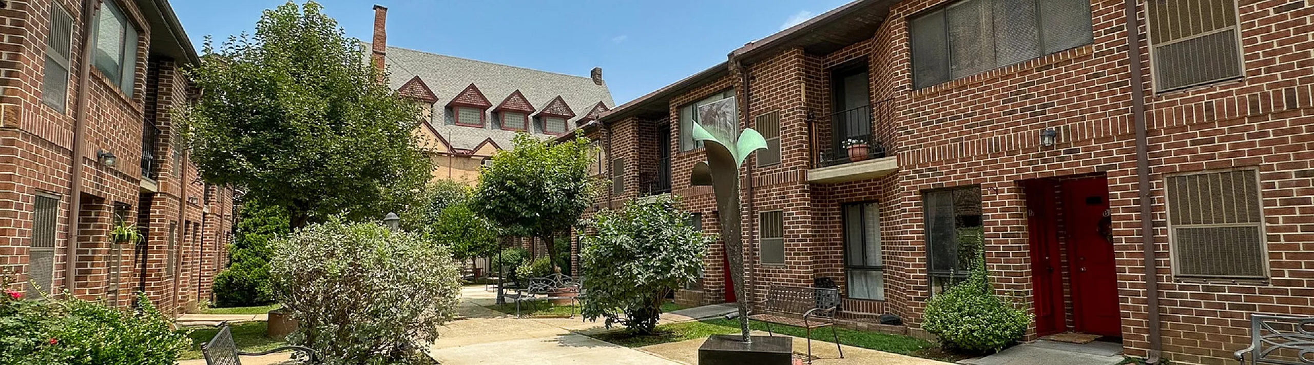 Charming courtyard view of a brick apartment complex with lush greenery, featuring cozy outdoor seating and vibrant red doors.