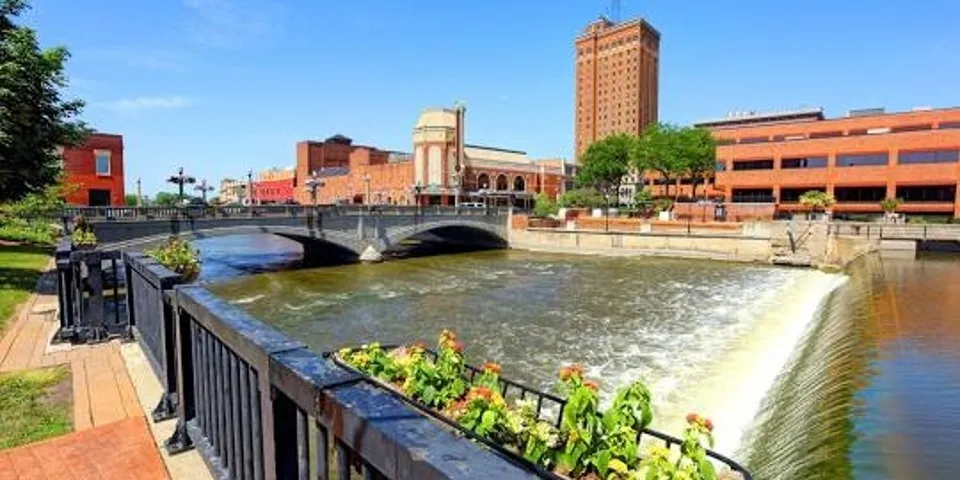 A large river with a waterfall flowing next to the property with brick buildings and an elegant bridge in the background.