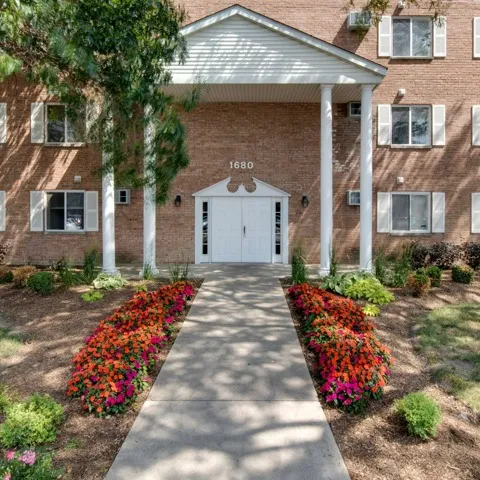 Main entrance to Colonial Village Apartments with four tall columns and bright flowers lining the walkway from a straight on perspective..