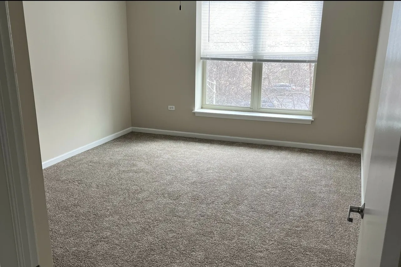 A second bedroom with grey carpeting, a large sunny window, and beige walls with white trim.