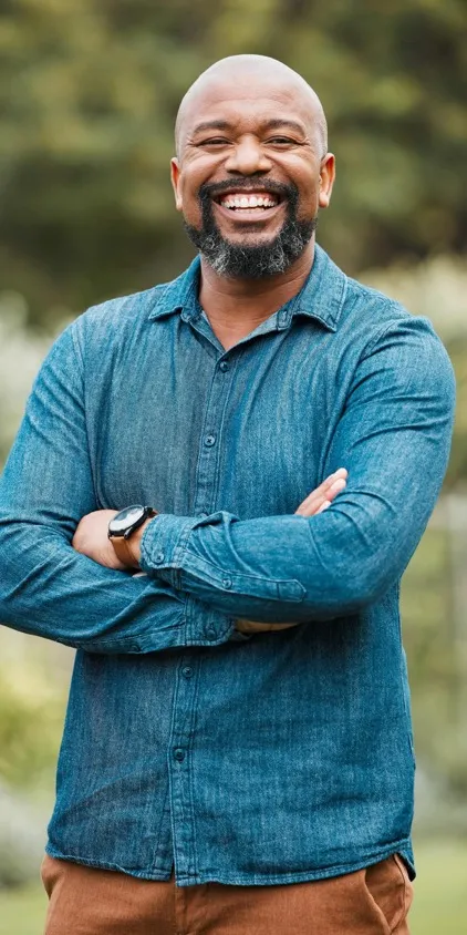 A smiling fifty five plus year old man in a blue shirt and brown pants with his arms folded.