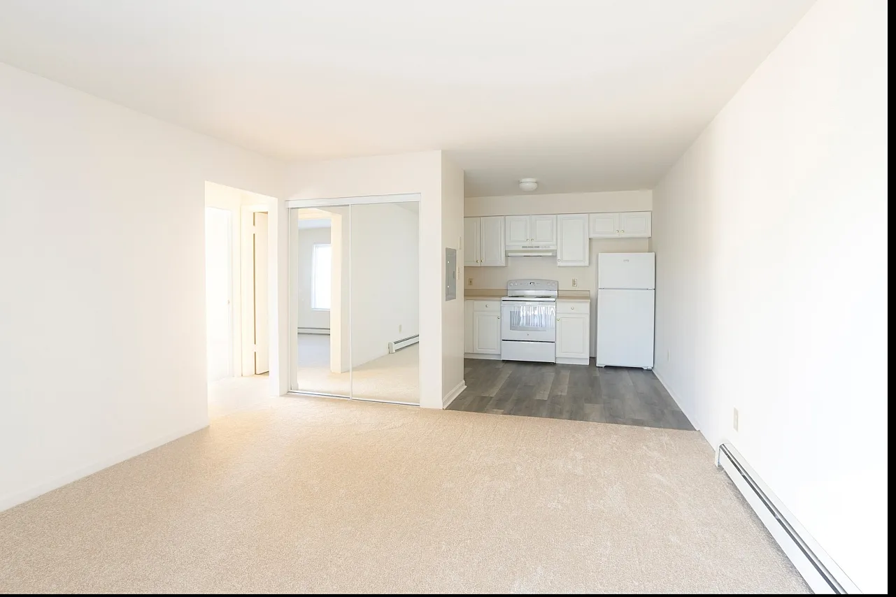 Open concept two bedroom apartment with a carpeted living room, off white walls, and white appliances.
