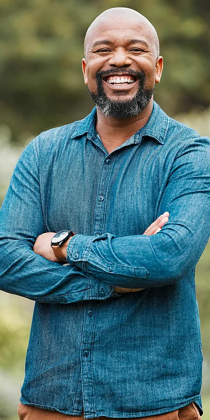 A smiling senior man in a blue shirt and brown pants with his arms folded.