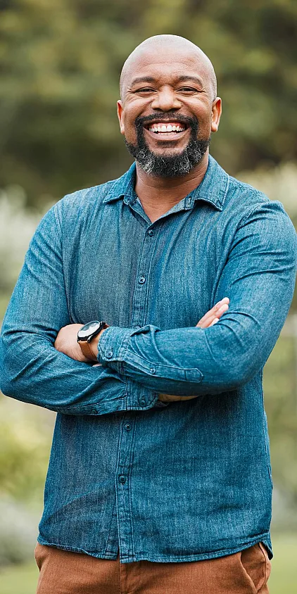 A smiling fifty five plus year old man in a blue shirt with his arms folded.