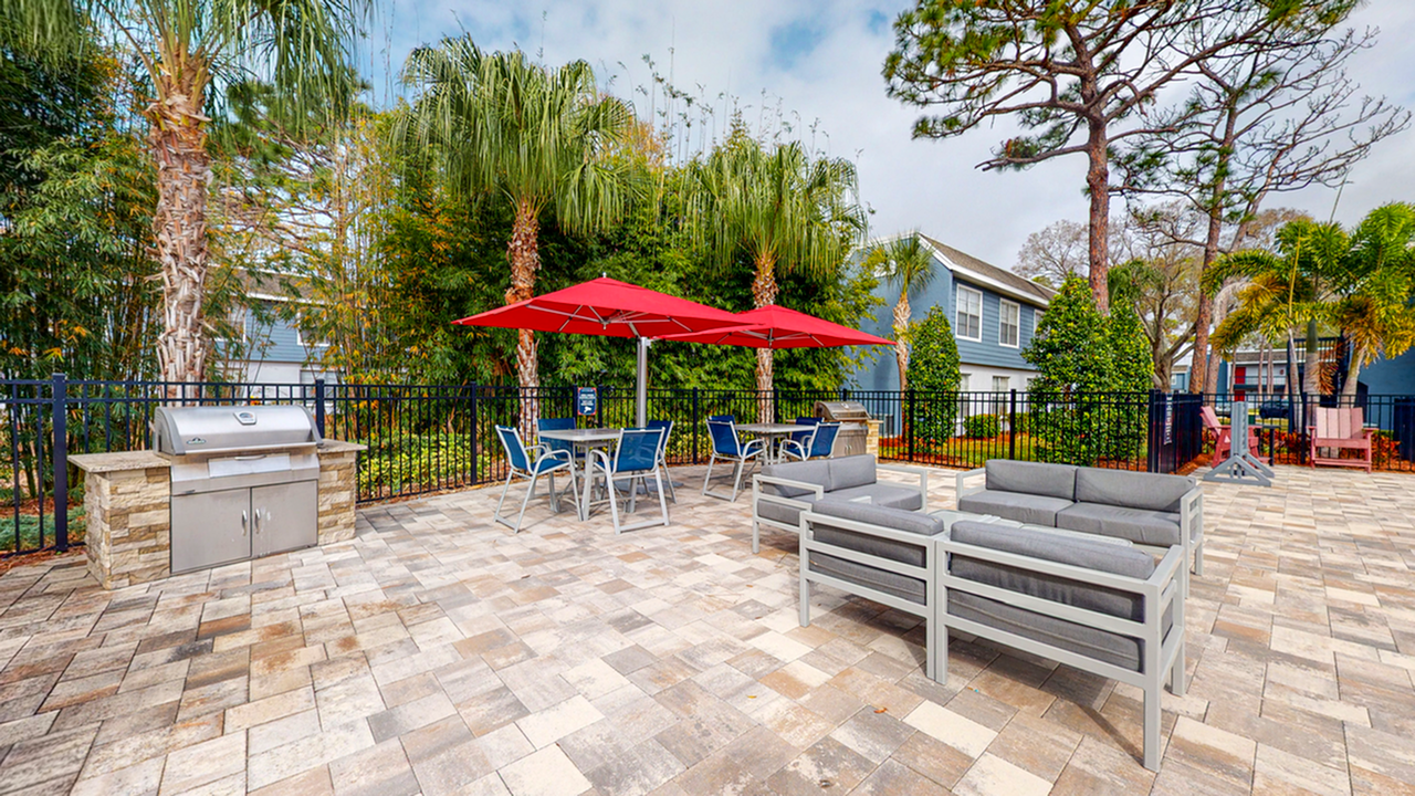 Pool grill area with cushioned gray patio furniture, gray table and blue chairs, red umbrellas, trees, blue apartments