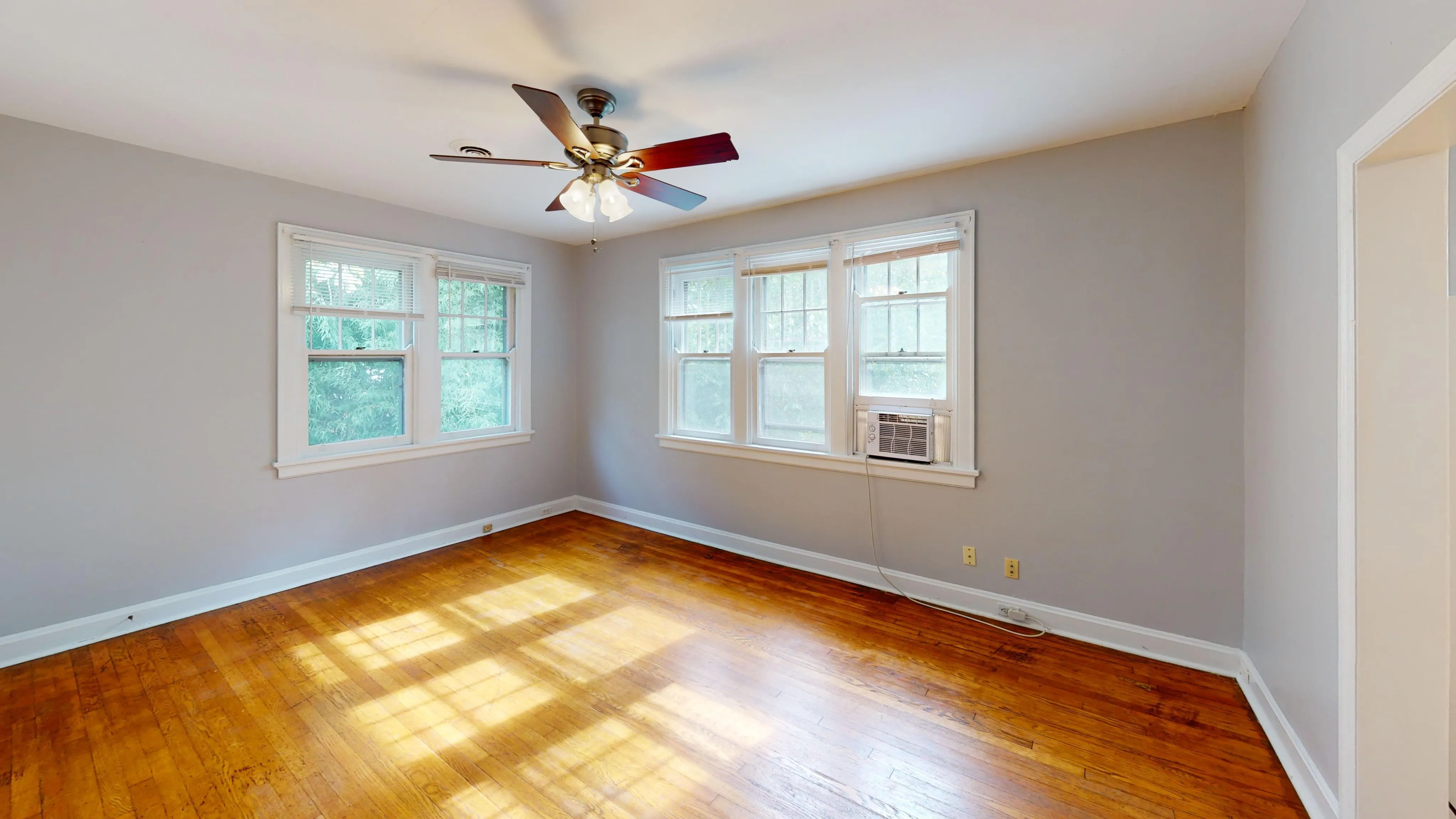 Sunlit corner room with hardwood floors and large windows featuring a ceiling fan and air conditioning unit.