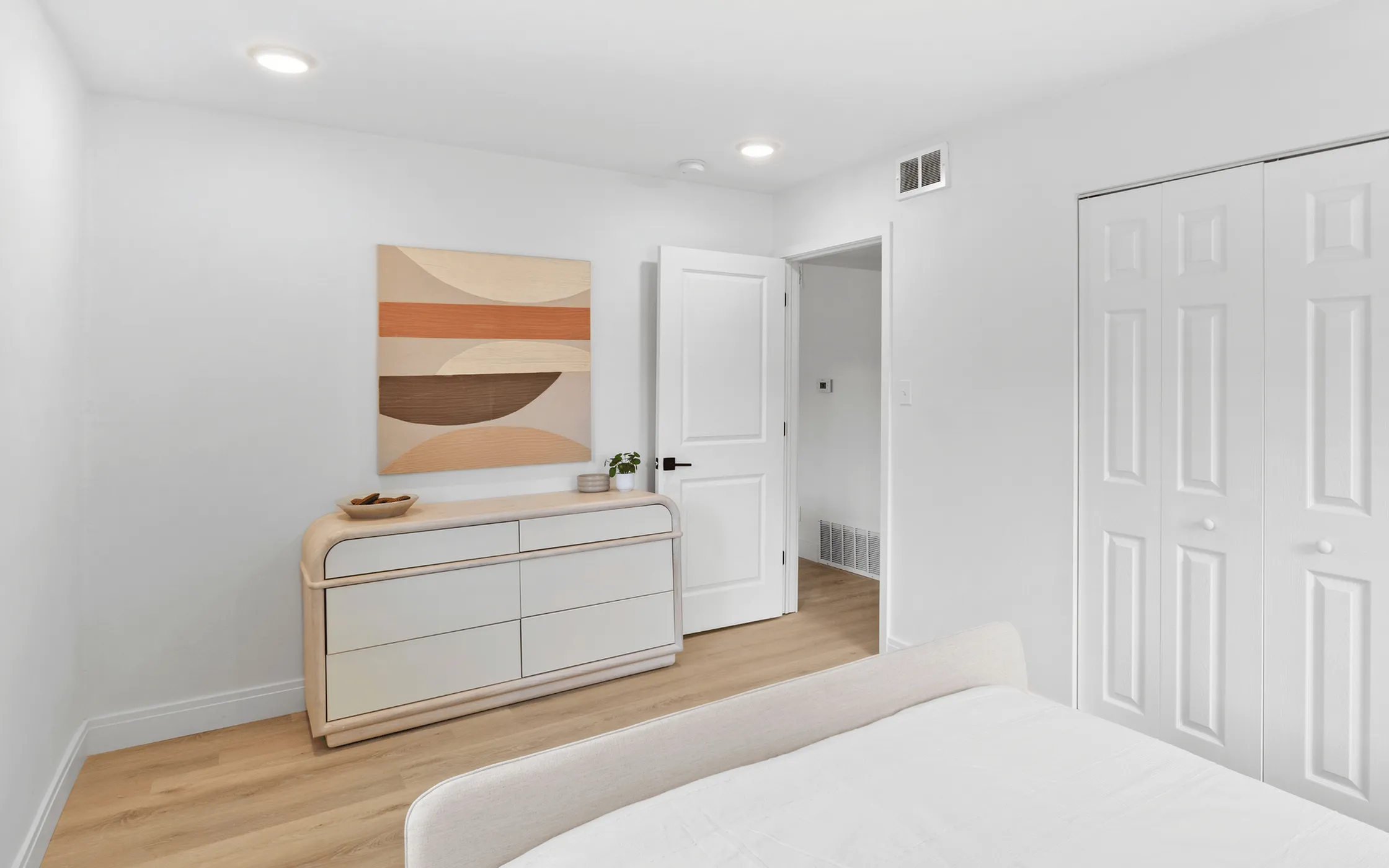 Bright and modern bedroom with minimalist decor, featuring a sleek dresser, abstract wall art, and spacious closet.