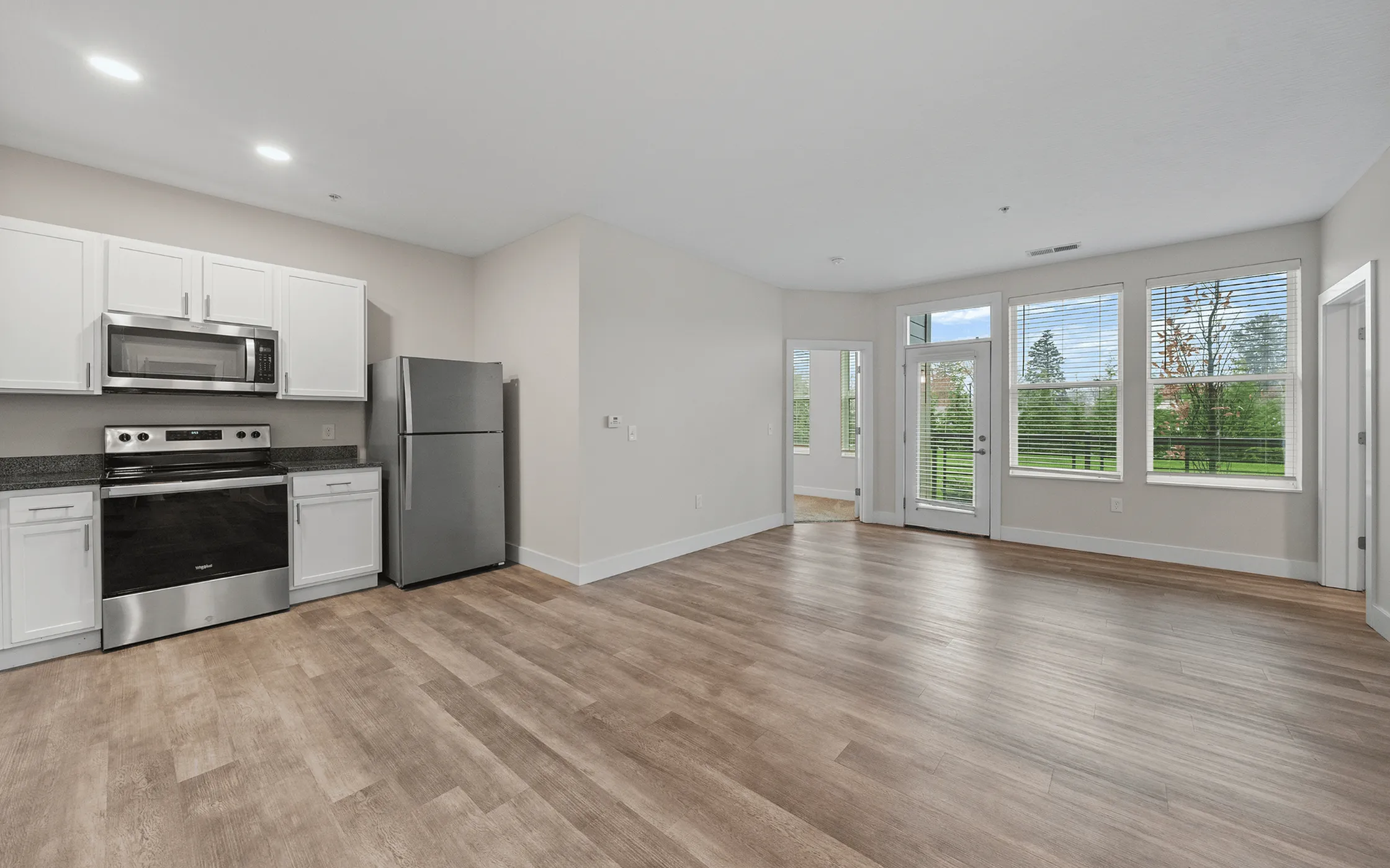 Bright open-concept kitchen and living area with sleek stainless steel appliances, hardwood floors, and large windows overlooking greenery.
