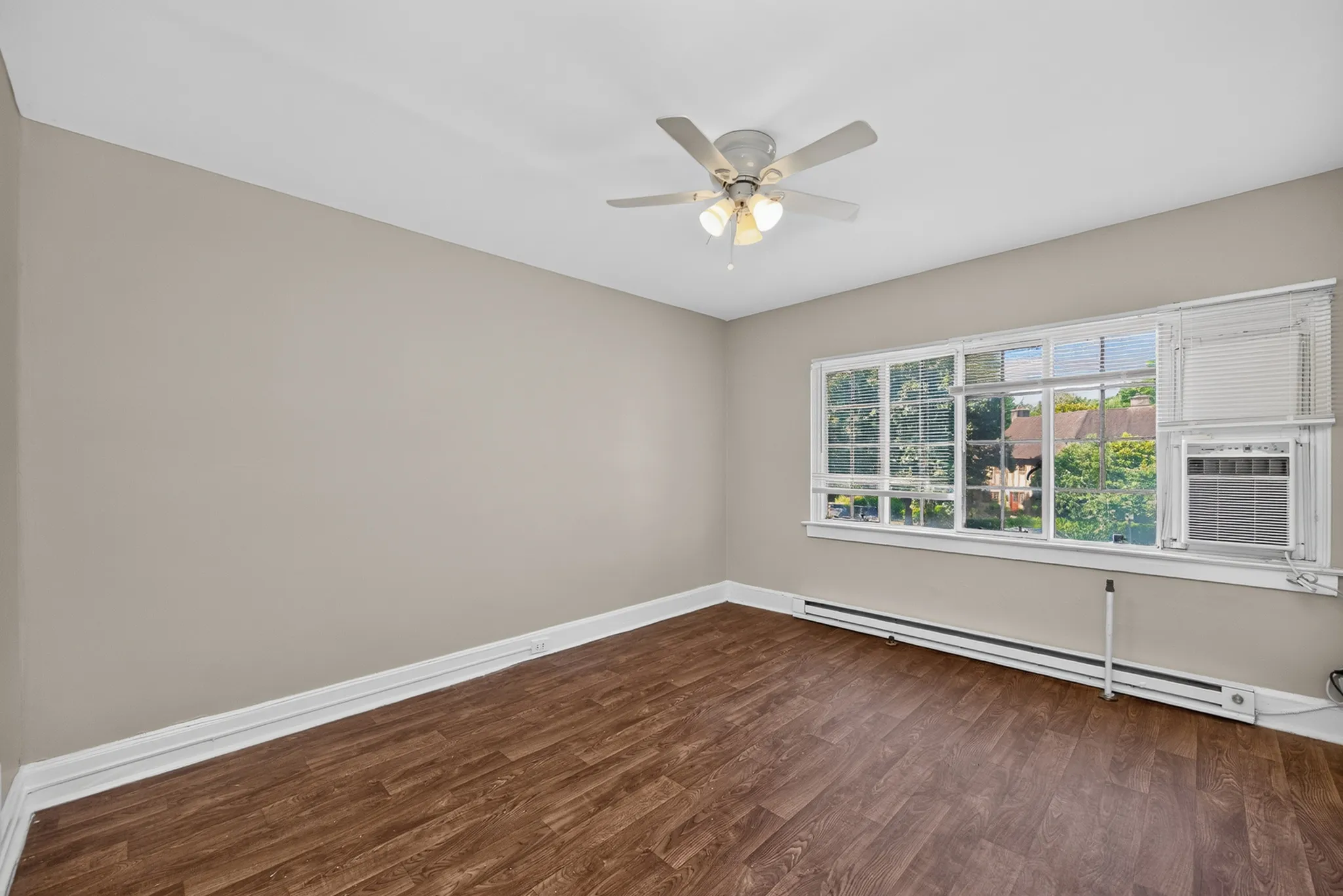 Bright, empty room with elegant wood flooring and a large window offering natural light, featuring a ceiling fan for optimal comfort.