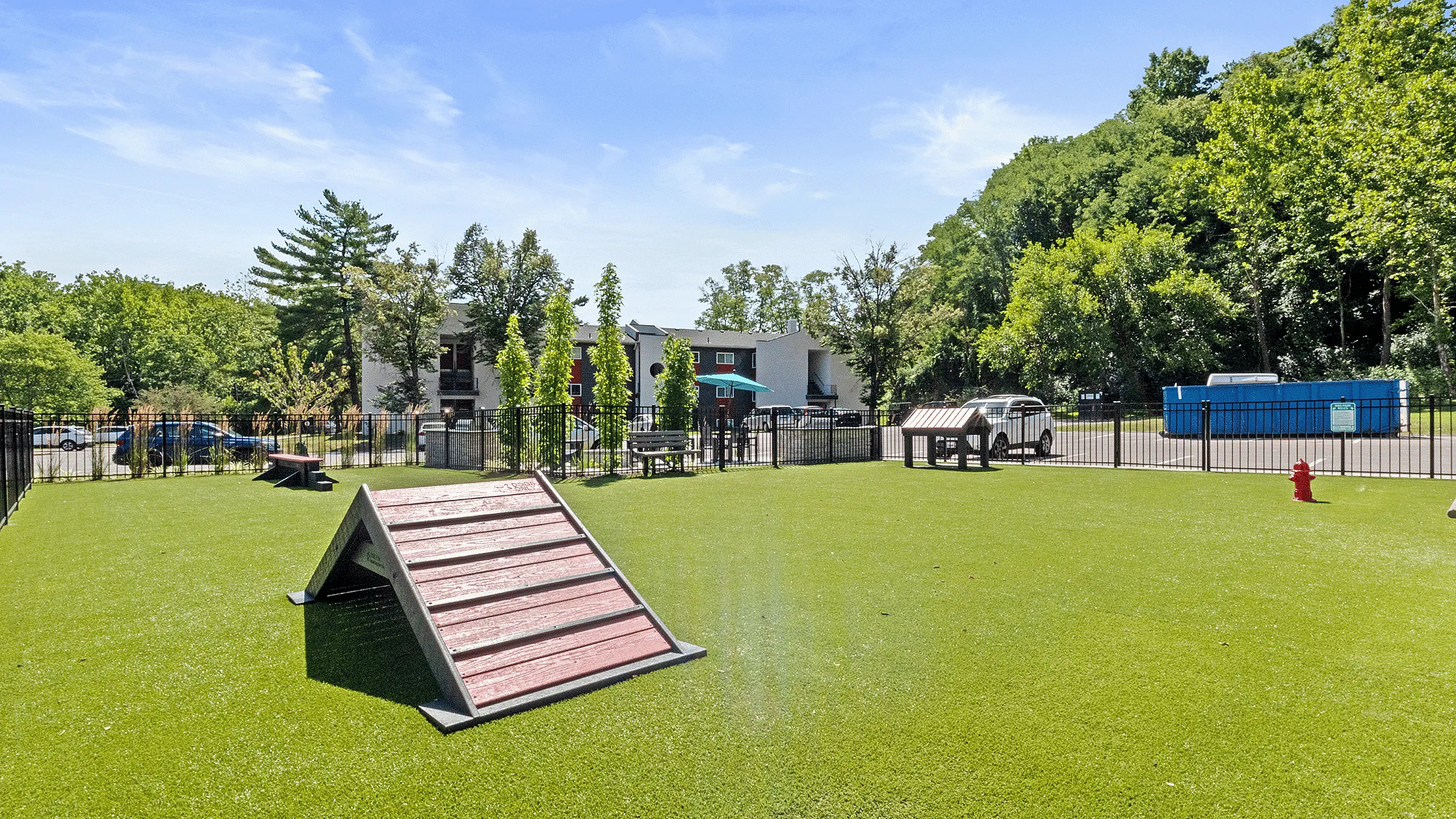 Charming dog park with agility equipment, lush green turf, and a scenic backdrop of trees and modern apartment buildings.