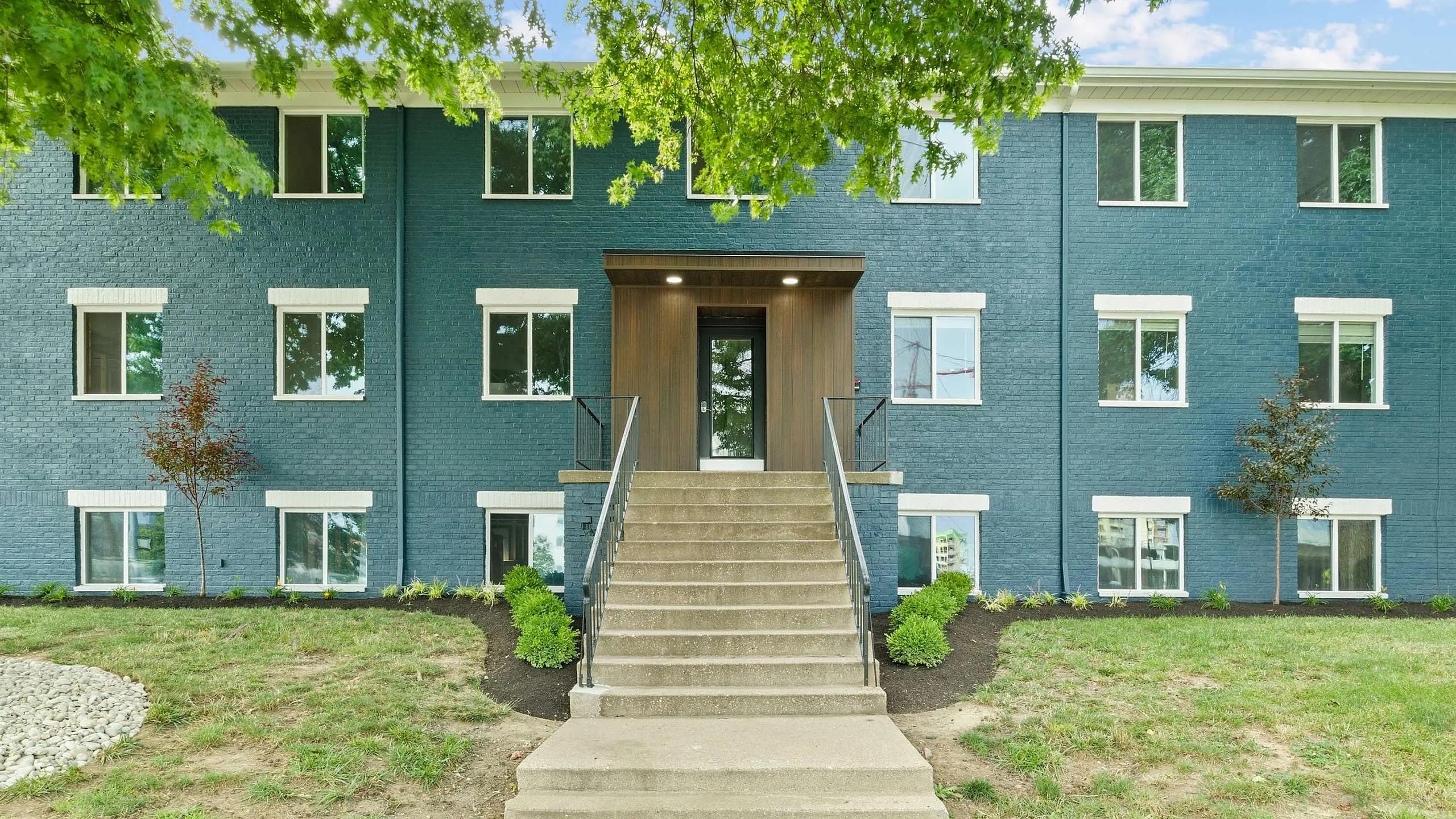 Charming apartment building with a modern blue brick exterior, lush landscaping, and a welcoming central entrance.
