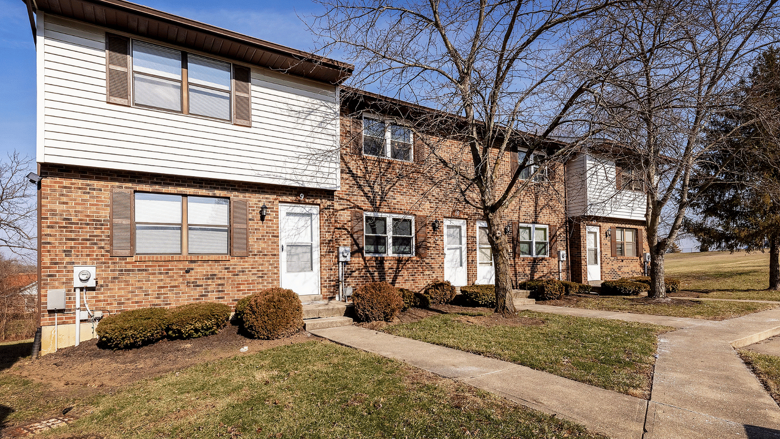 Fairfield Square Apartments Exterior, Charming brick townhouse with modern design, featuring large windows and vibrant green landscaping under a clear blue sky.