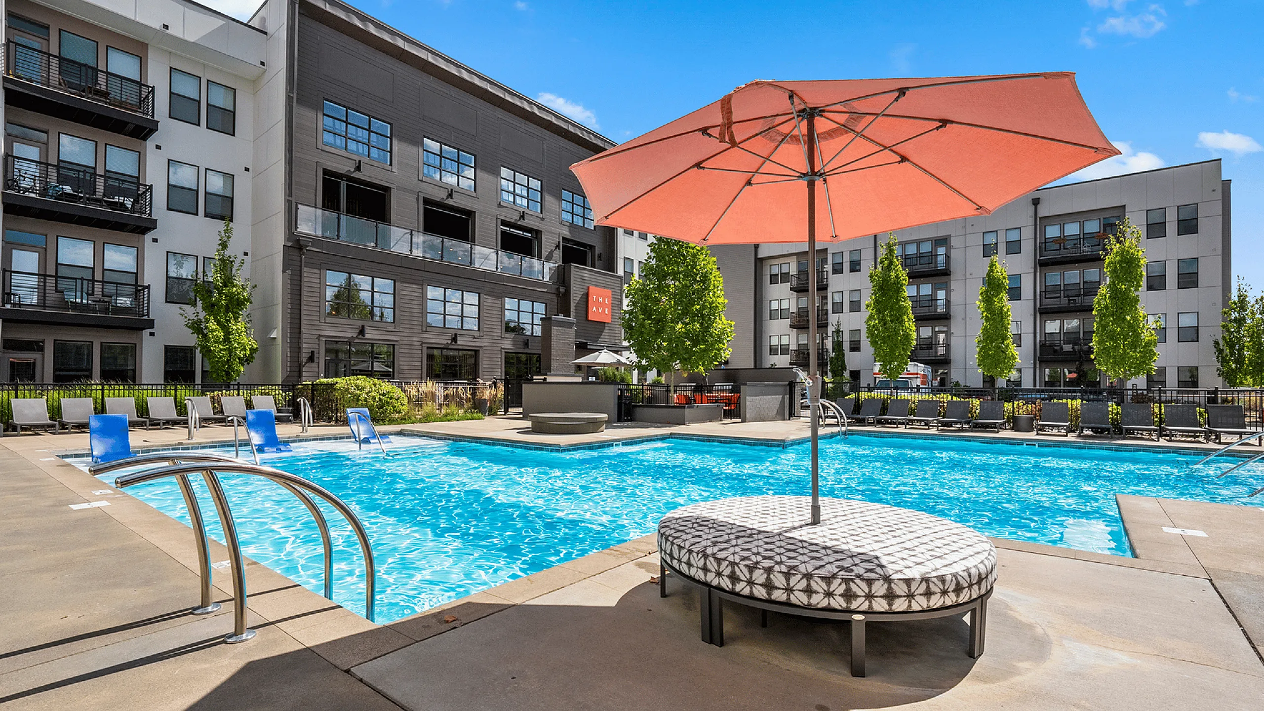 Modern apartment complex with inviting pool, sun loungers, and vibrant orange umbrella, perfect for relaxation and leisure under a clear blue sky. The Ave apartments in Clintonville, The Ave apartments in columbus ohio, columbus ohio apartm