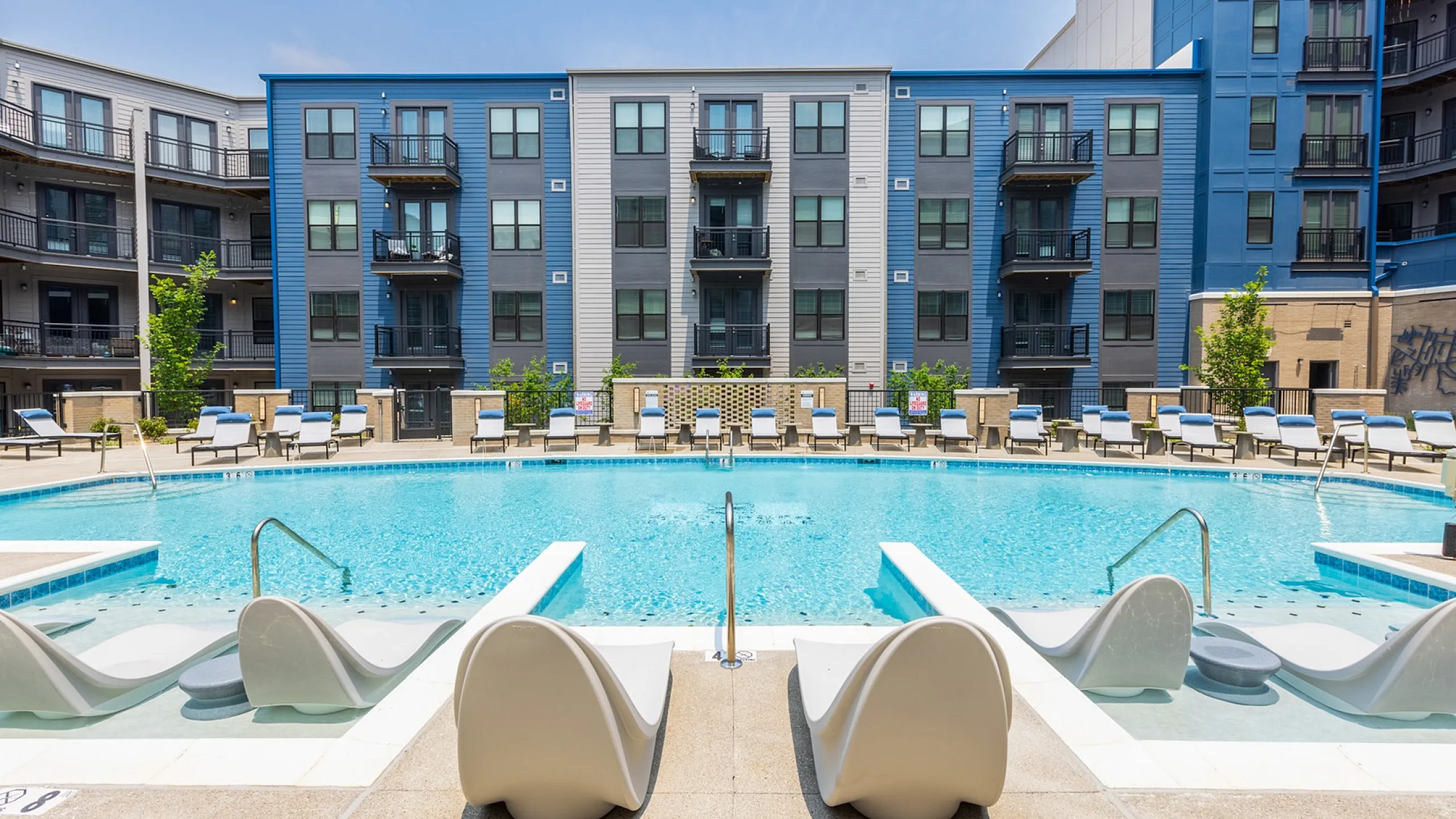 Modern apartment complex with a stunning resort-style pool, surrounded by sun loungers and vibrant landscaping.
