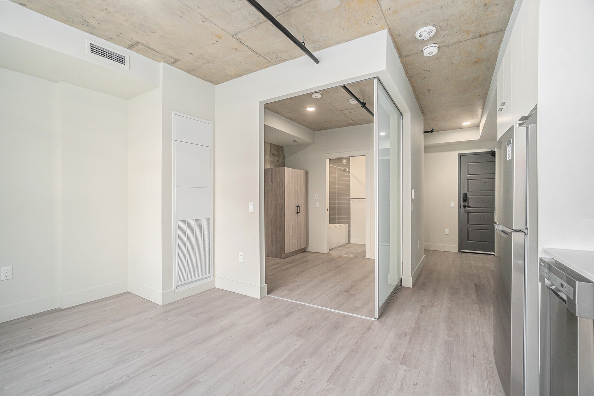 Modern apartment interior with sleek light wood flooring, exposed concrete ceiling, and stainless steel appliances featuring sliding glass doors leading to a spacious bathroom and storage area.