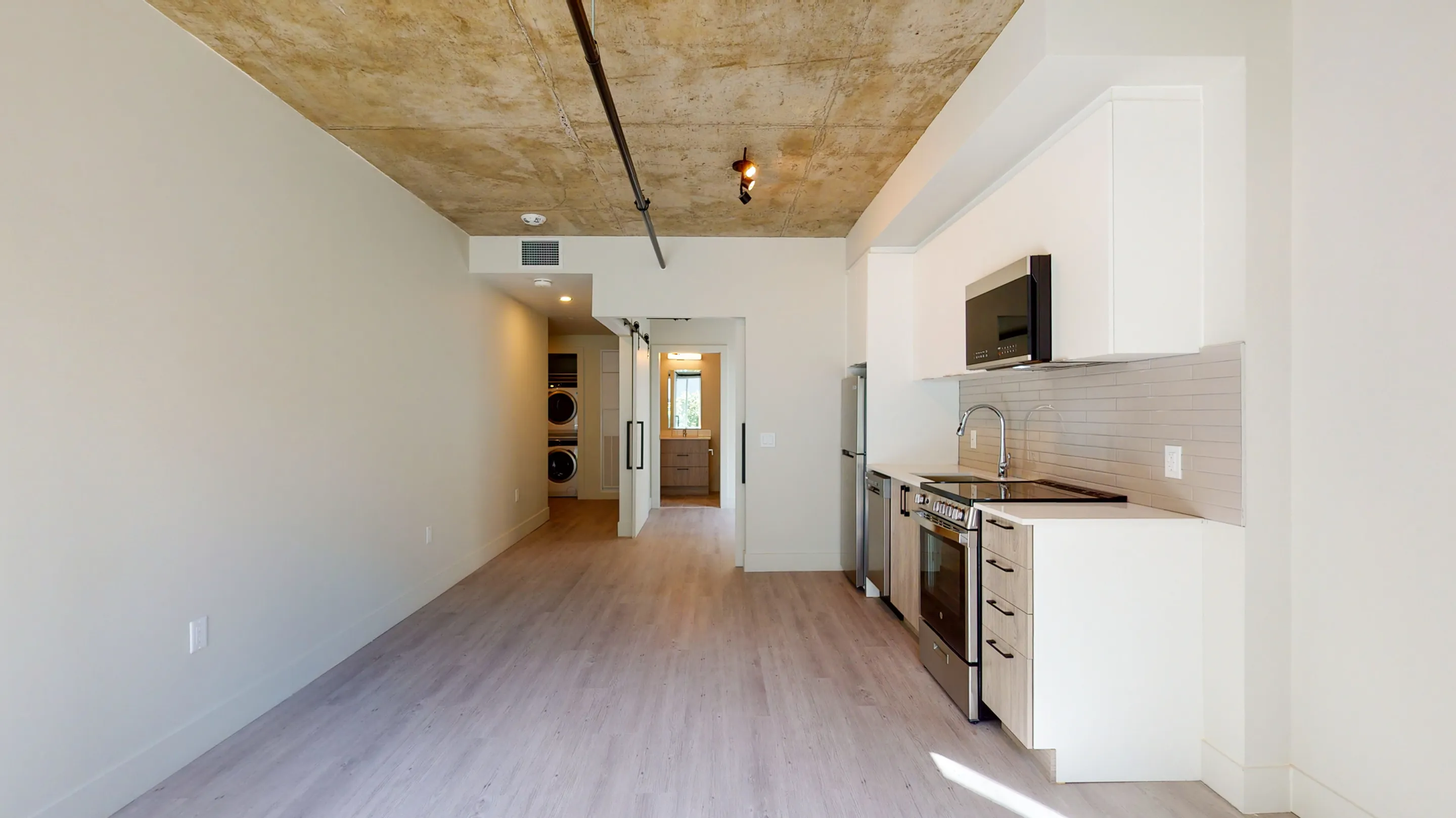Modern open-concept kitchen with sleek white cabinetry, stainless steel appliances, and natural wood flooring, leading to a bright hallway and laundry area.