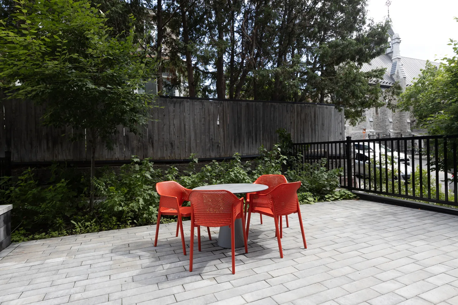 Charming outdoor patio with vibrant red chairs and a cozy table, surrounded by lush greenery and a modern fence.