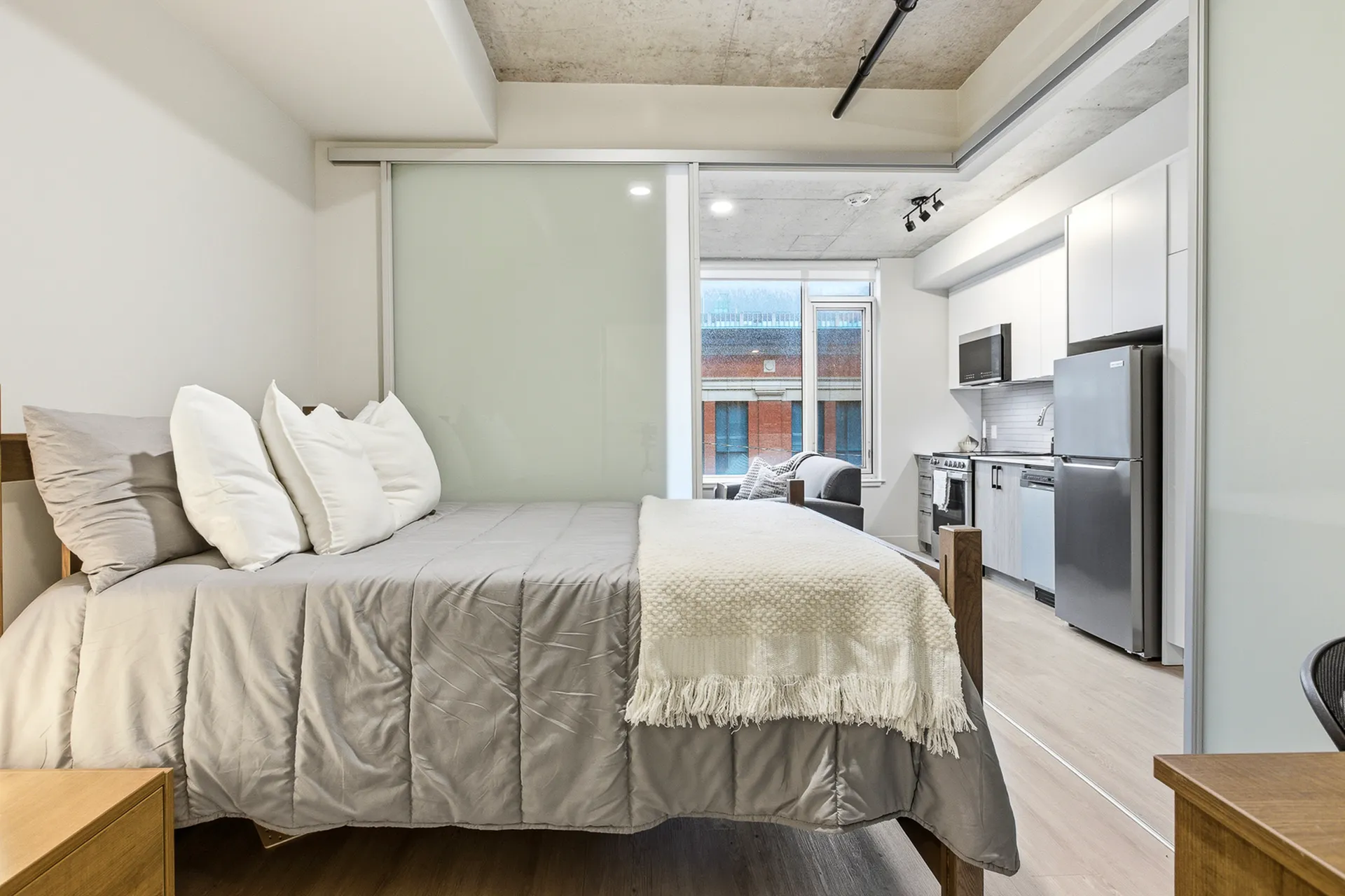 Modern studio apartment with sleek design, featuring a cozy bed adjacent to an open kitchen with contemporary appliances and a view of an urban building through the window.