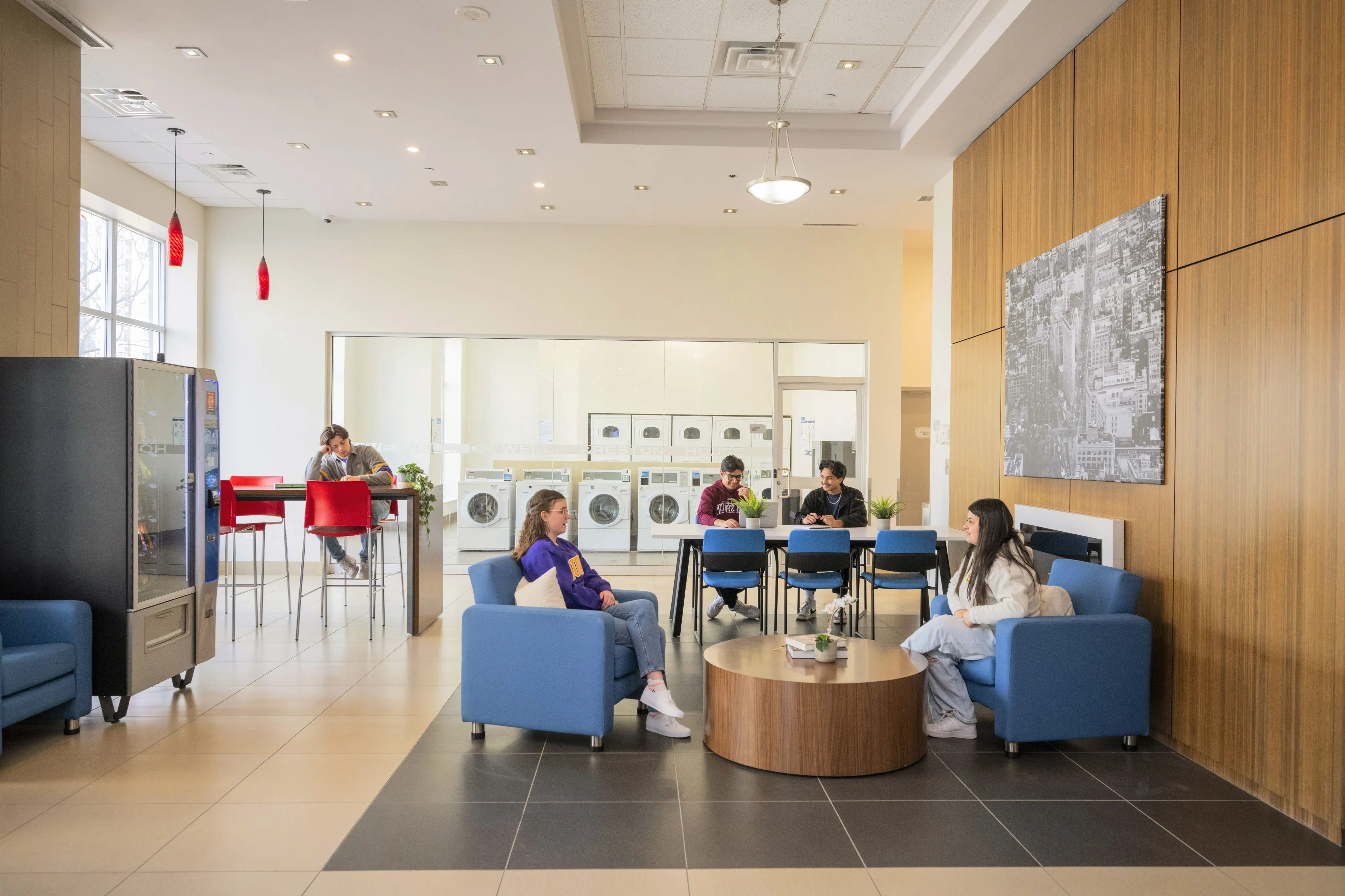 Bright and modern community lounge with comfortable seating, vibrant red accents, and a row of washing machines in the background.