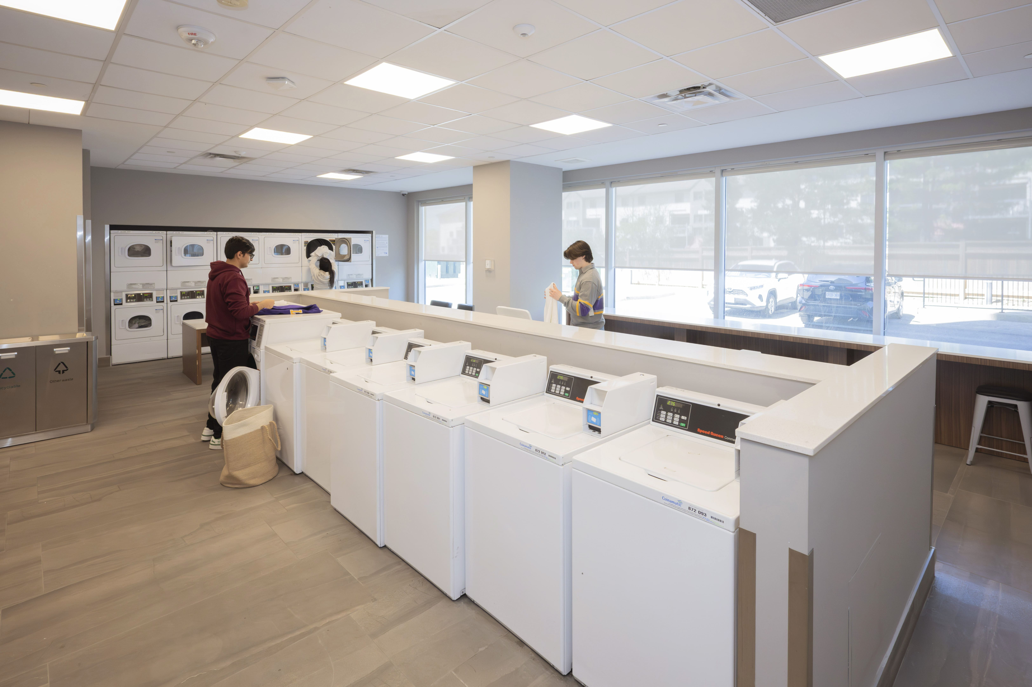Modern communal laundry room with multiple washing and drying machines, bright and spacious design, featuring large windows and a comfortable environment.