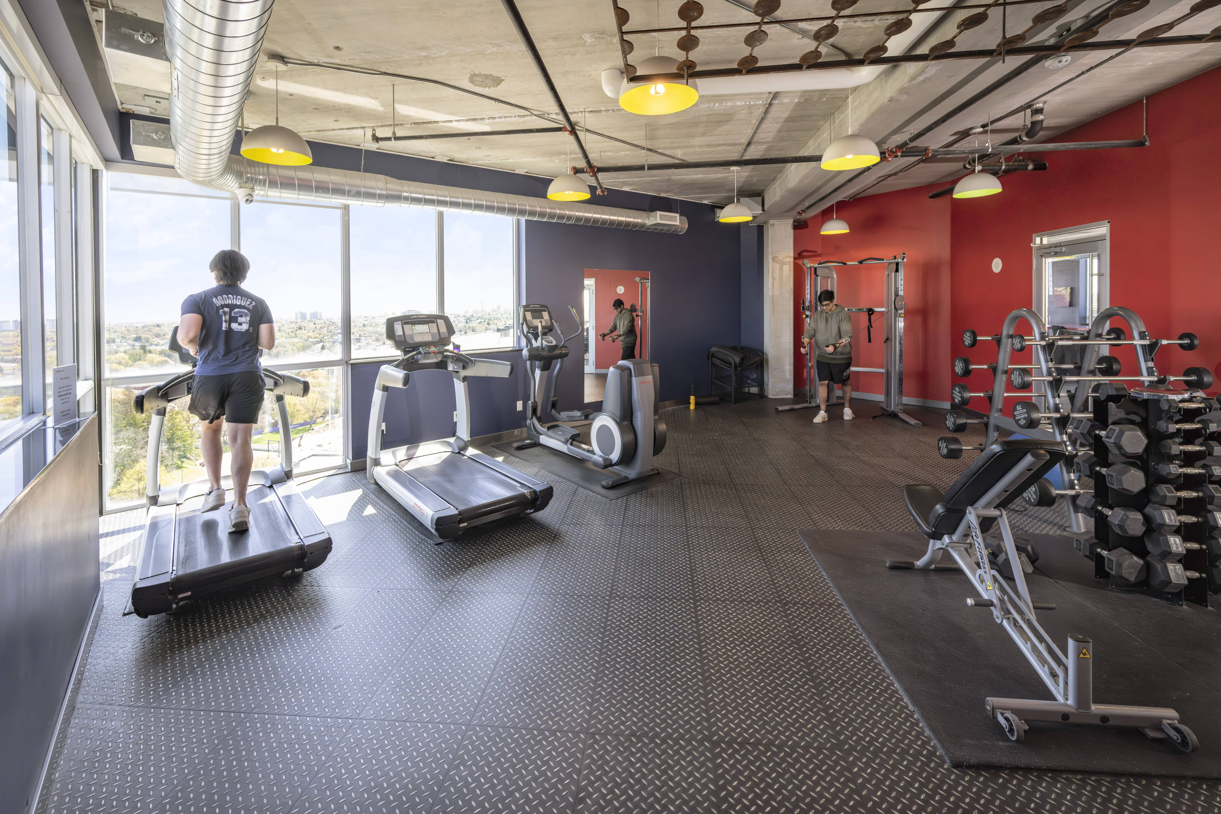 Bright and modern gym with panoramic views, featuring treadmills, an exercise bike, free weights, and red accent walls.