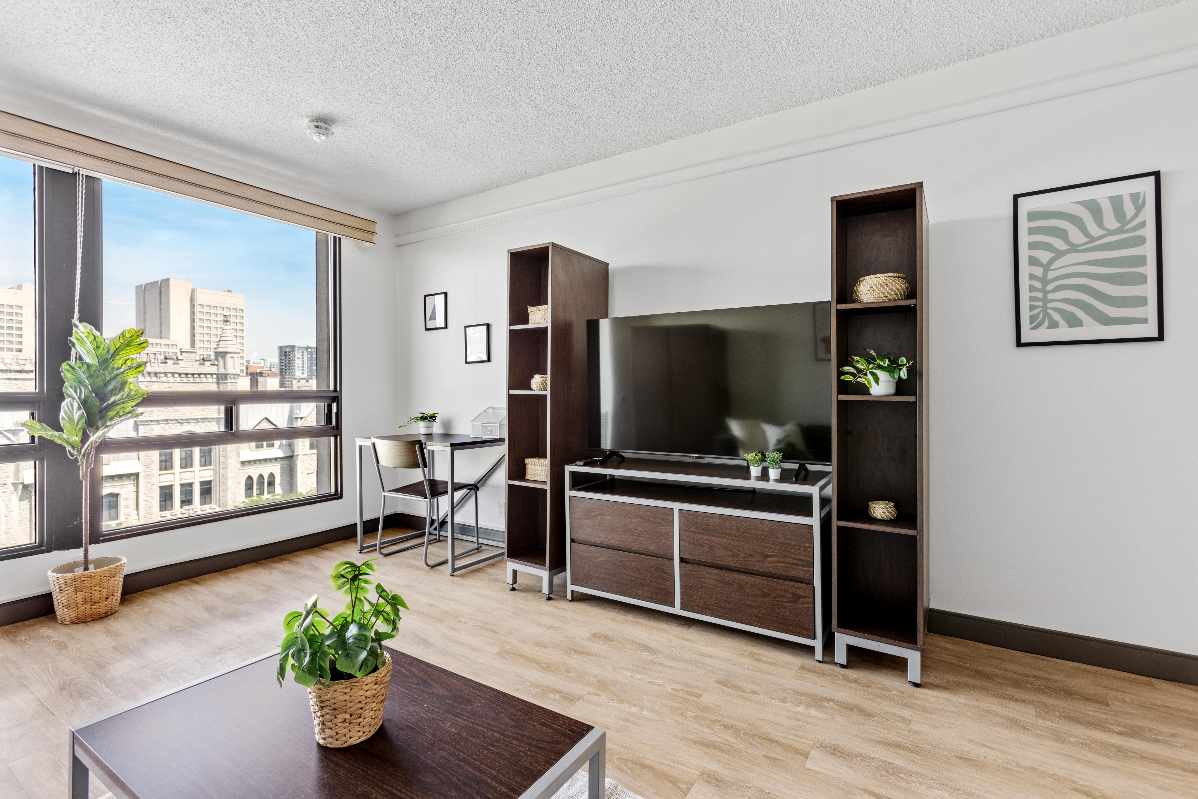 Bright and modern living room with large windows offering city views, sleek wooden furniture, and vibrant green plants.