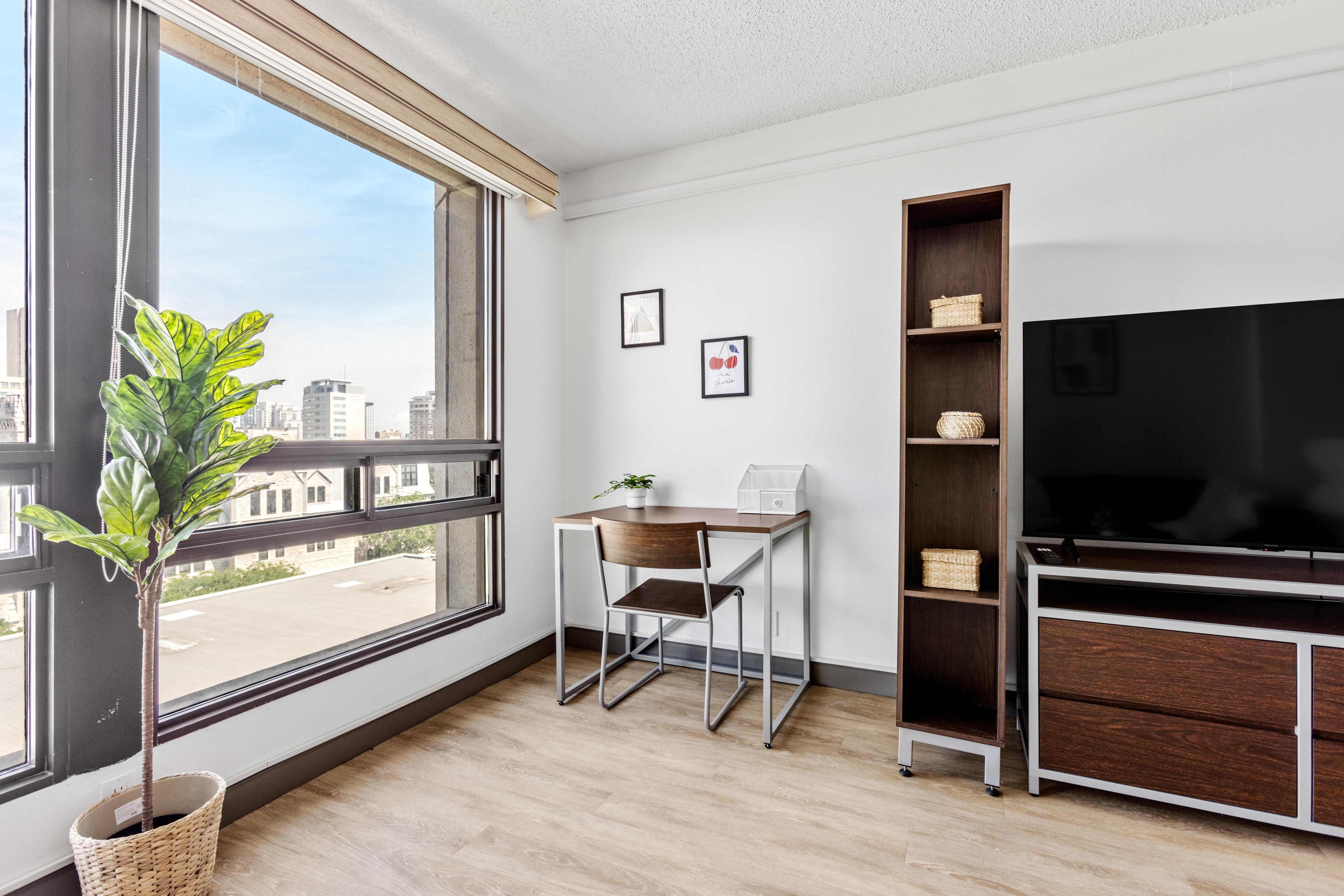 Elegant city apartment room with large windows offering abundant natural light, featuring a stylish desk area, modern shelving unit, and sleek television setup.