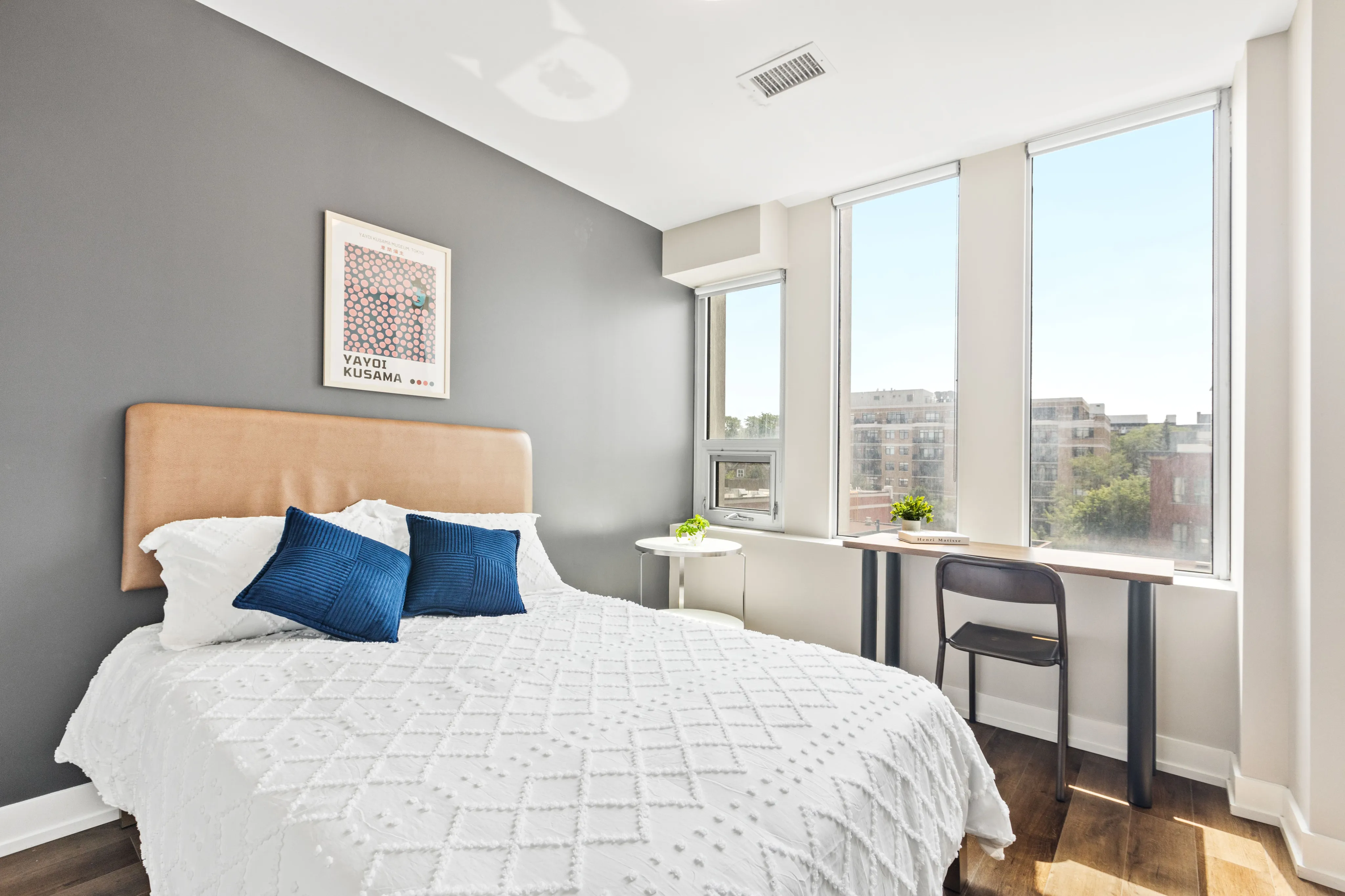 Bright and modern bedroom with large windows, stylish decor, and a cozy bed, offering a serene urban view.
