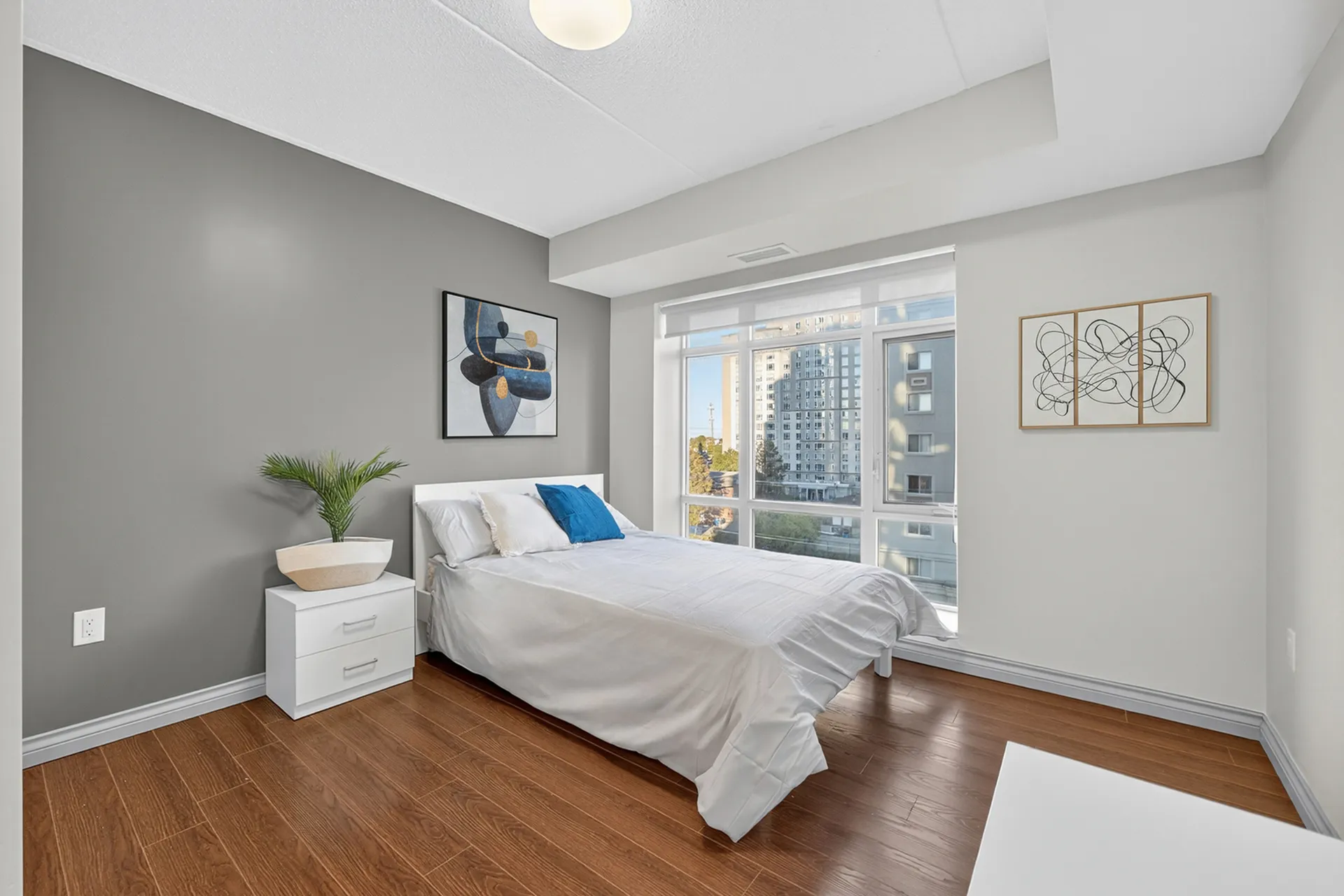 Bright and modern bedroom with hardwood floors, featuring a cozy bed and large windows offering a city view.