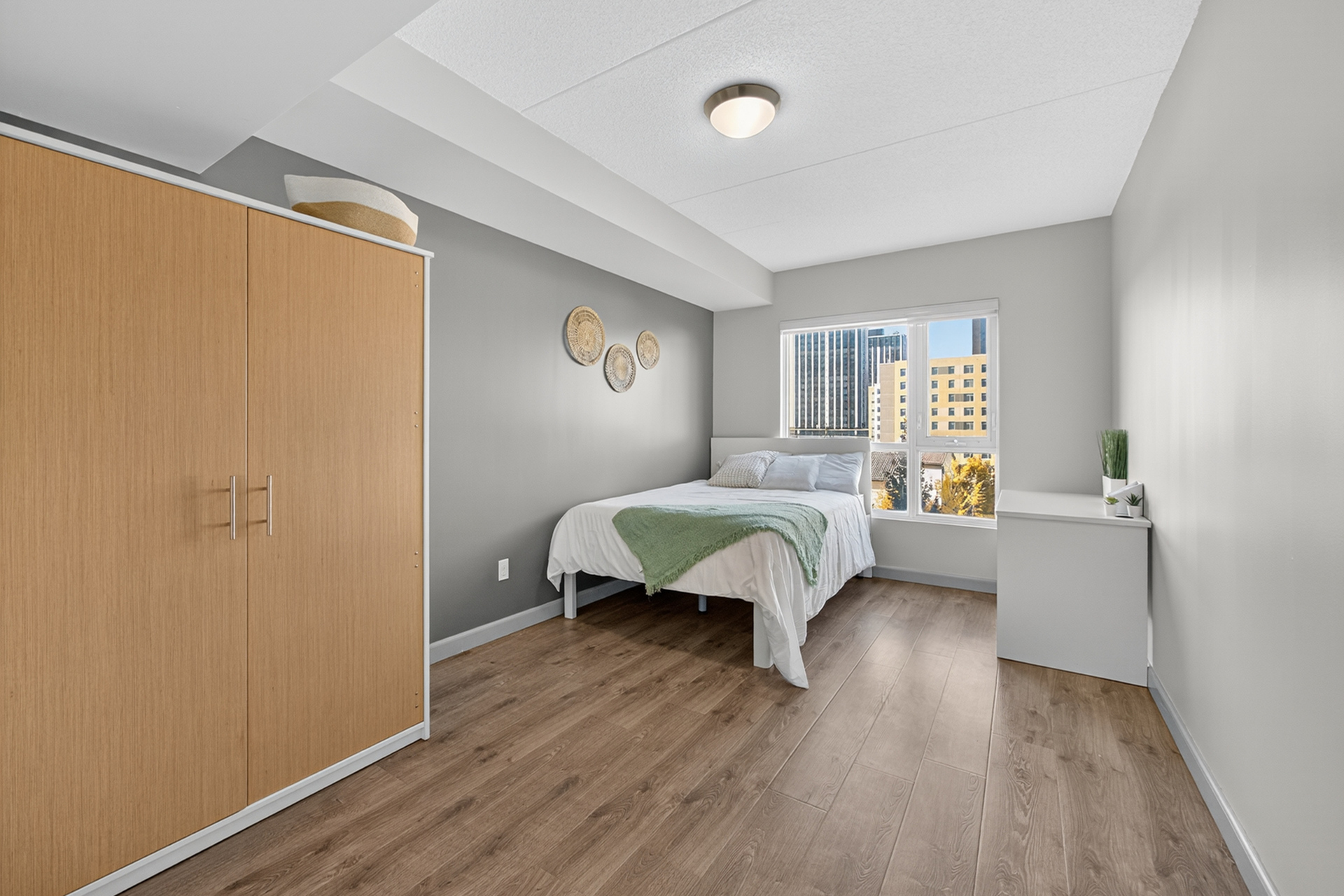 Bright, modern bedroom with sleek wood flooring, featuring a cozy bed, large window with city view, and stylish decor.