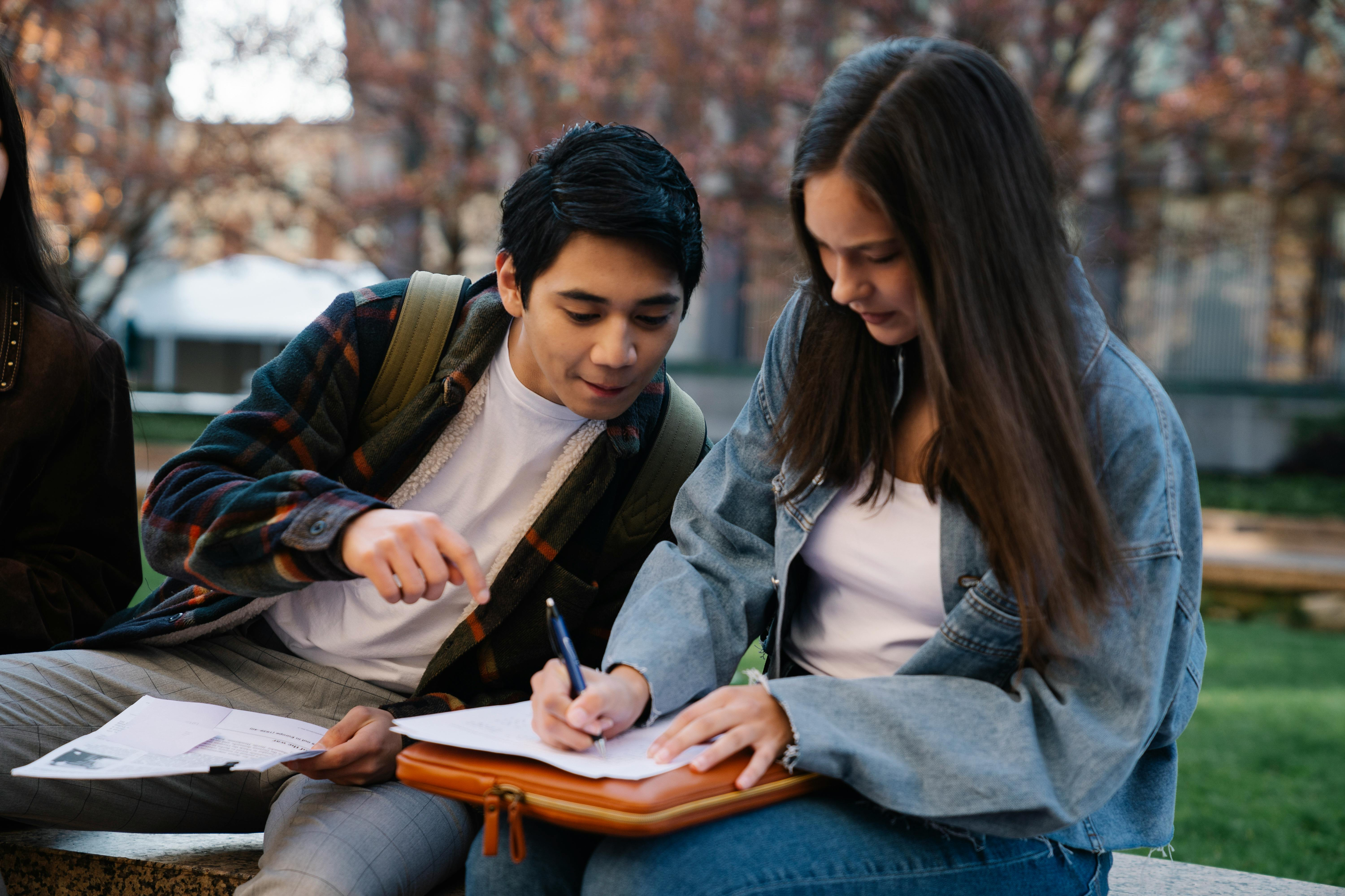 Two students working together outdoors on a pleasant day, with trees and buildings in the background, fostering a collaborative learning environment.
