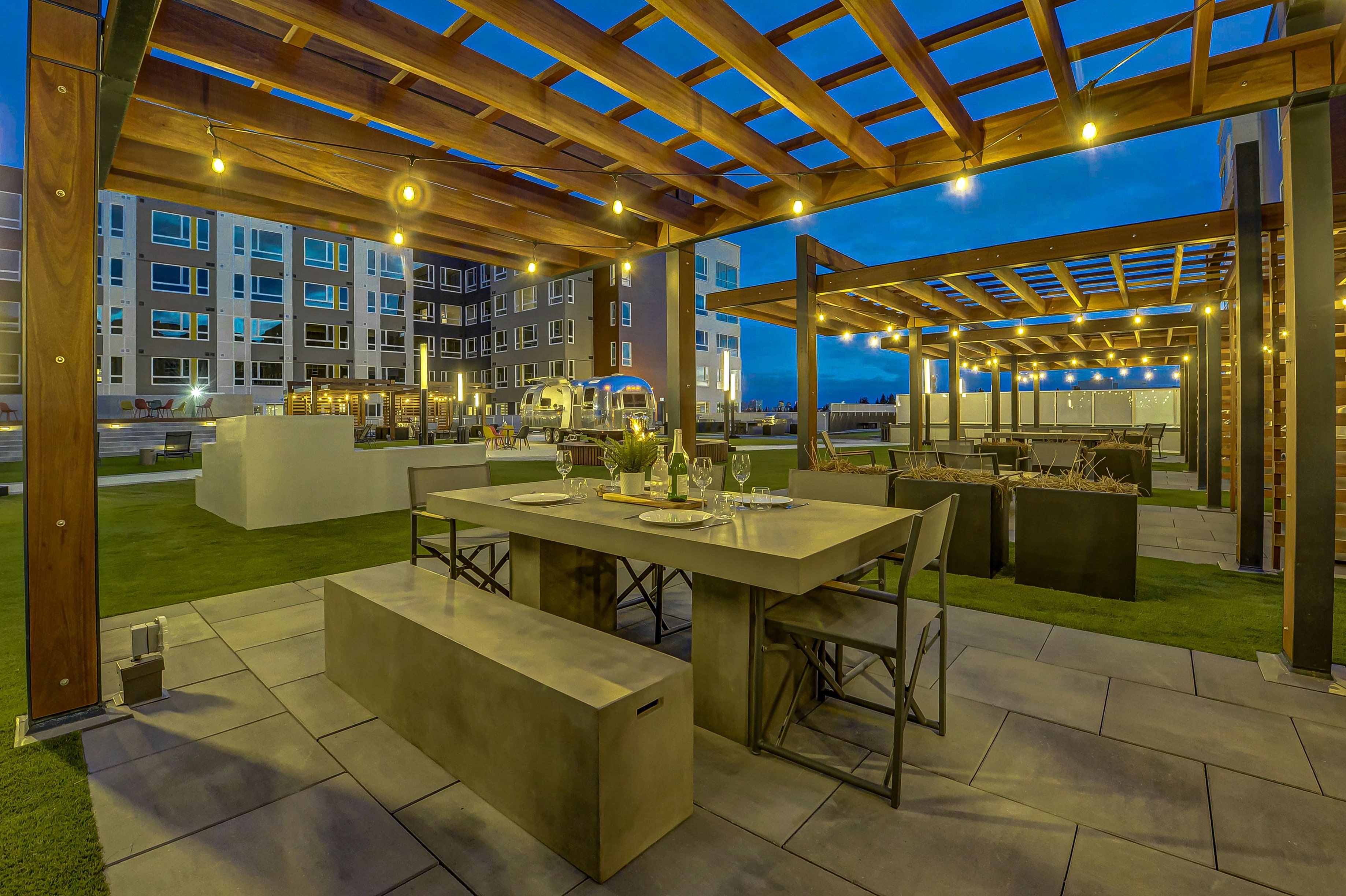 Chic outdoor dining area with modern concrete table, stylish seating, and ambient string lights under a wooden pergola, set against a modern apartment backdrop at dusk.