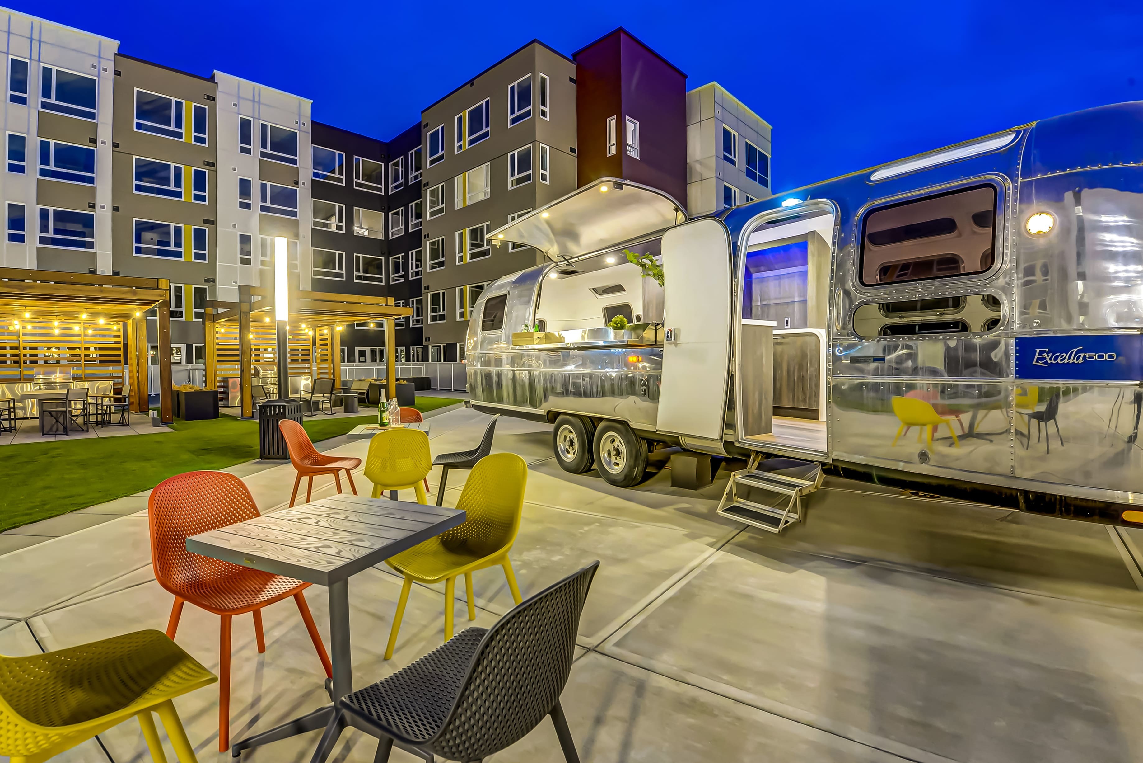 Vibrant urban apartment complex with chic outdoor seating and retro Airstream bar, perfect for social gatherings under the stars.