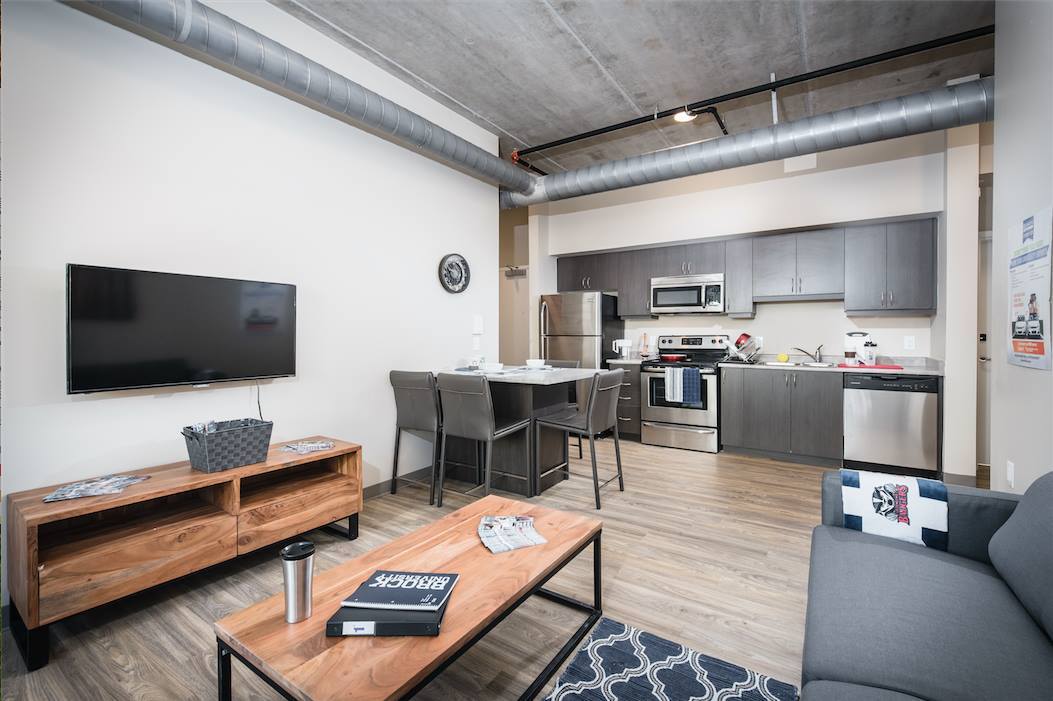 Modern open-concept living room and kitchen with sleek stainless steel appliances, cozy seating, and stylish wooden accents under industrial-style exposed ductwork.