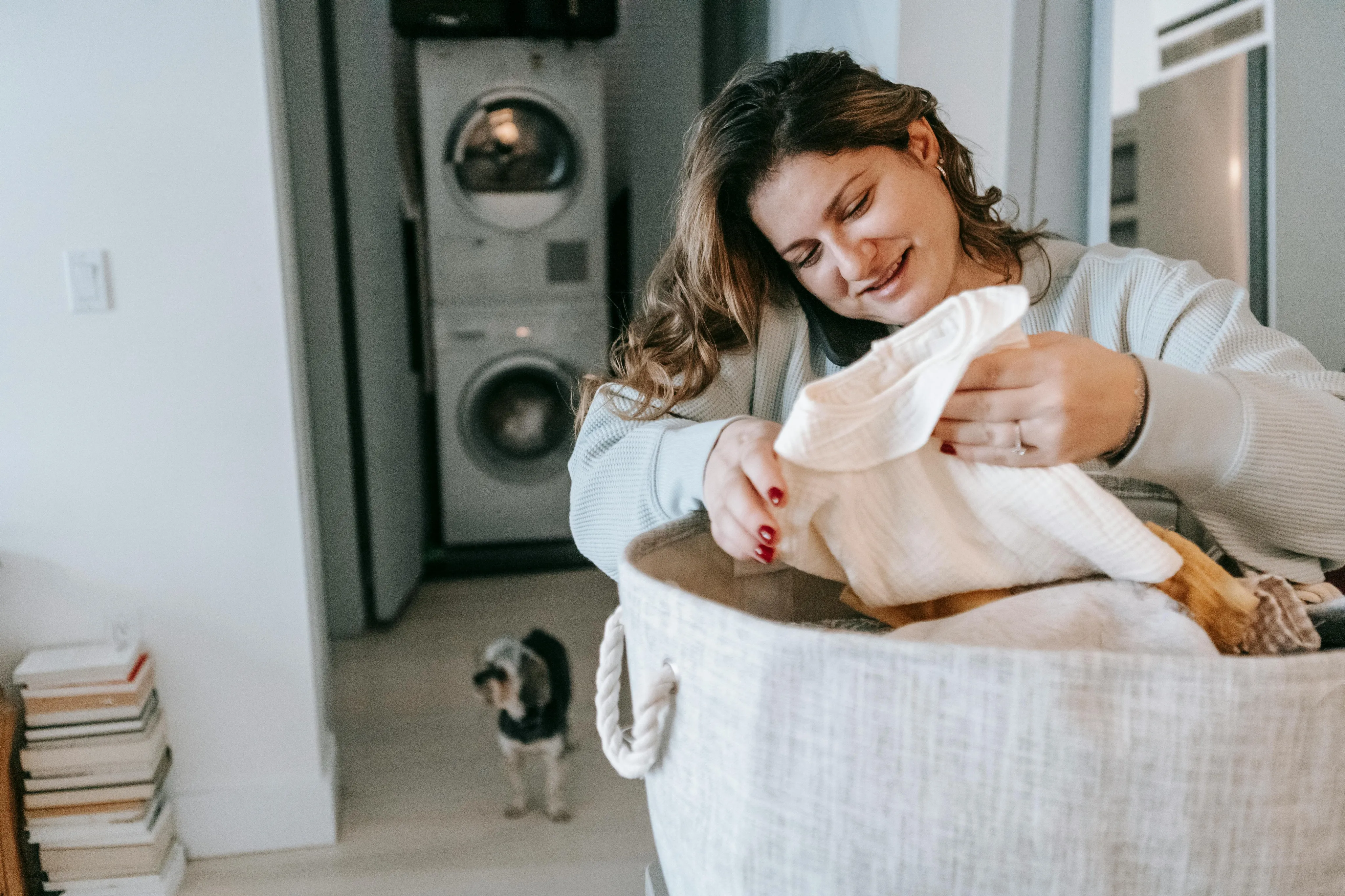 Cozy laundry area in a modern apartment with a smiling woman folding clothes and a washer-dryer unit in the background; inviting lifestyle scene with a curious dog.